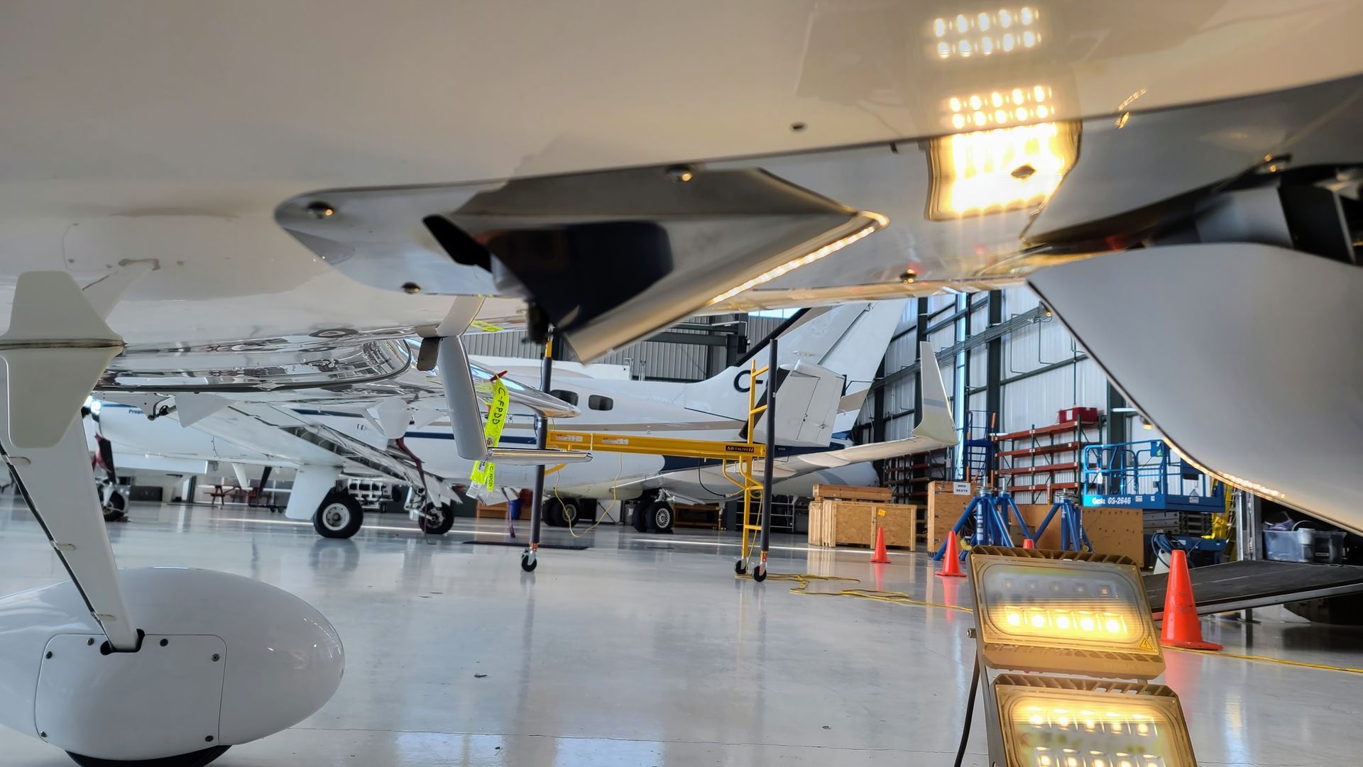 A plane's wing is visible. Other aircraft and equipment are in the background. Bright lights illuminate the interior.