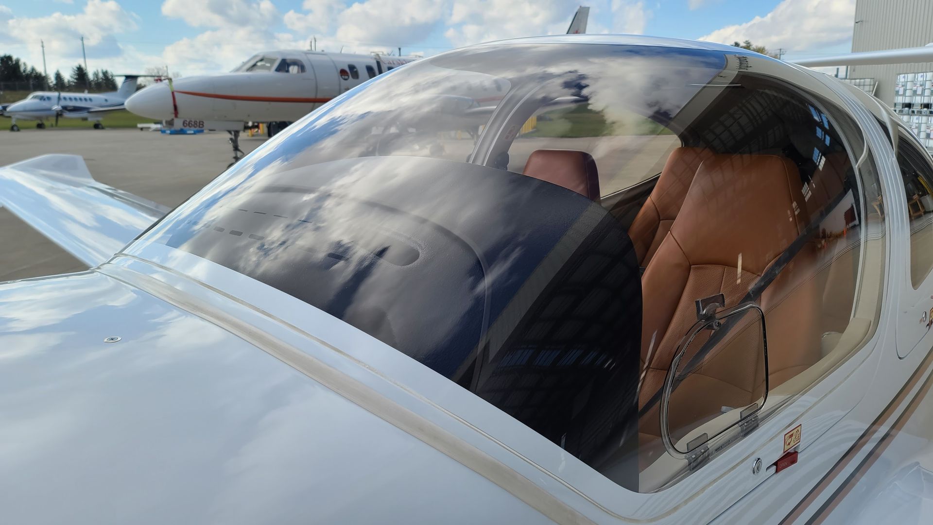 White airplane cockpit with clear windshield, tan seats. Another plane in the background on tarmac.