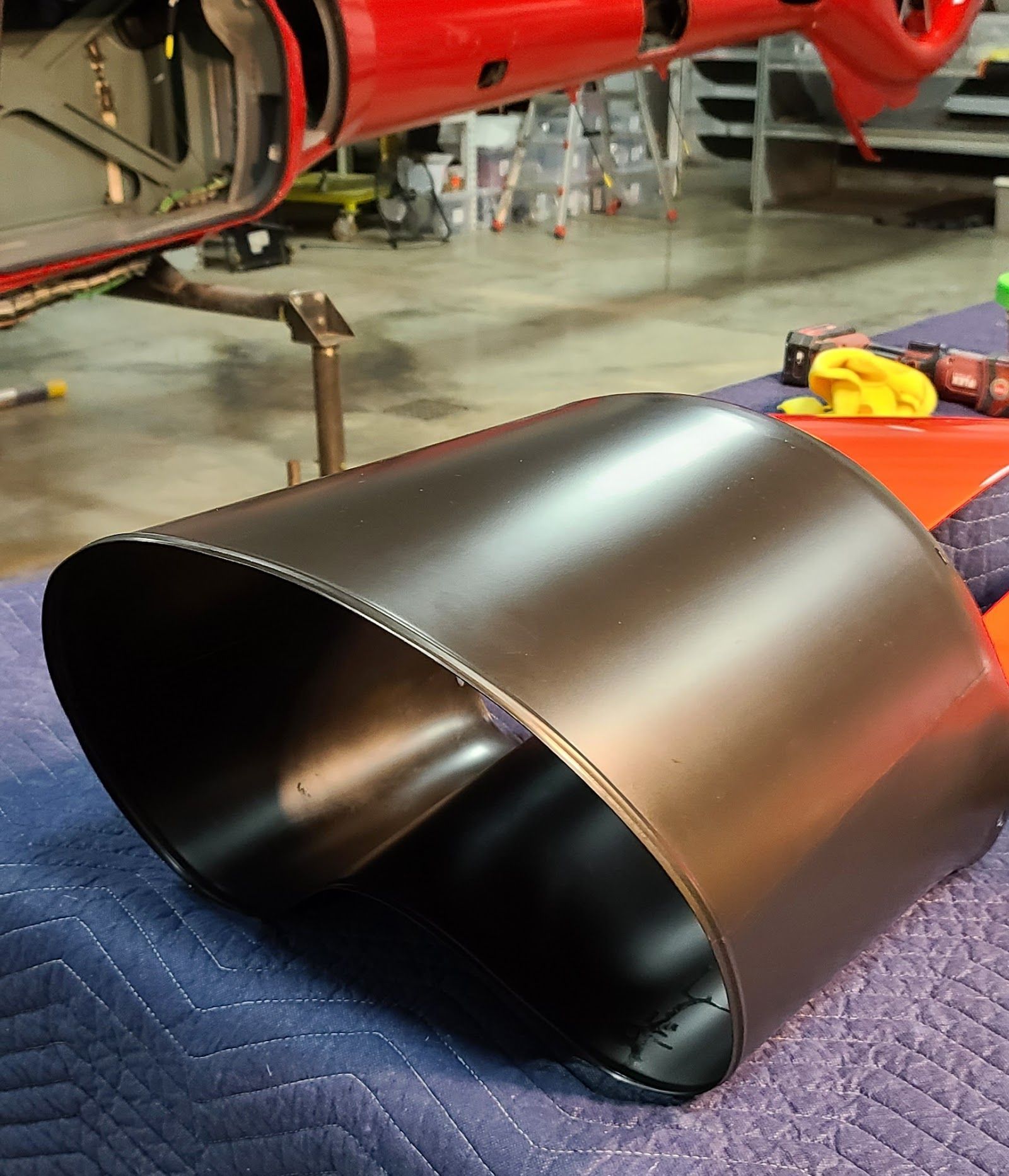 Black, oval-shaped exhaust pipe on a blue surface, with a red car in the background.