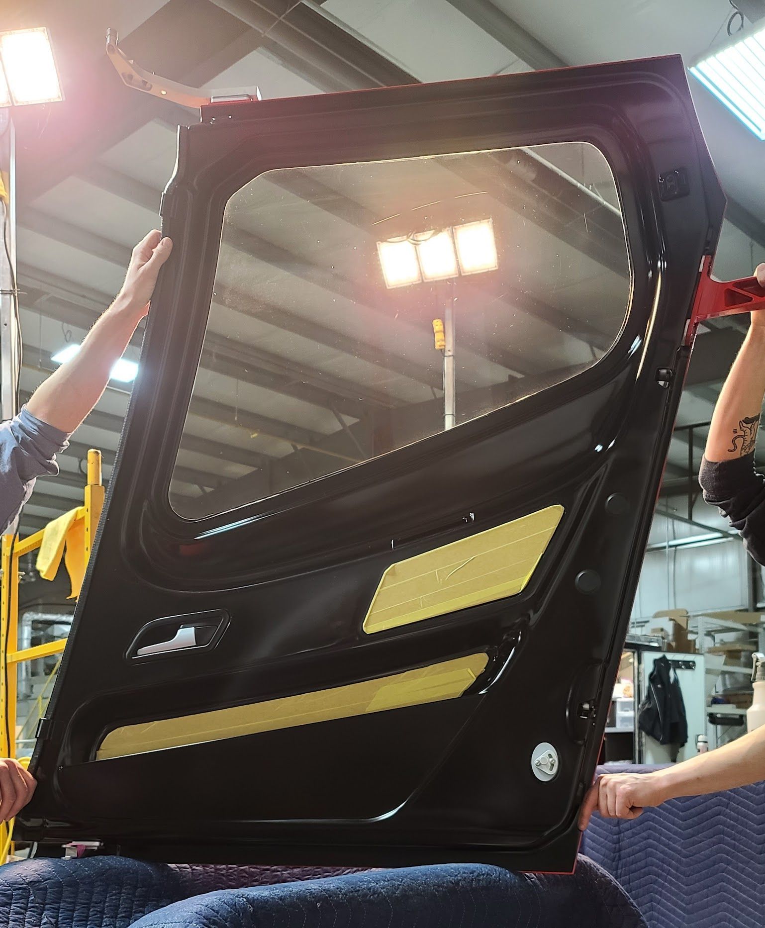 People holding a black car door with a window, indoors, lit by studio lights.