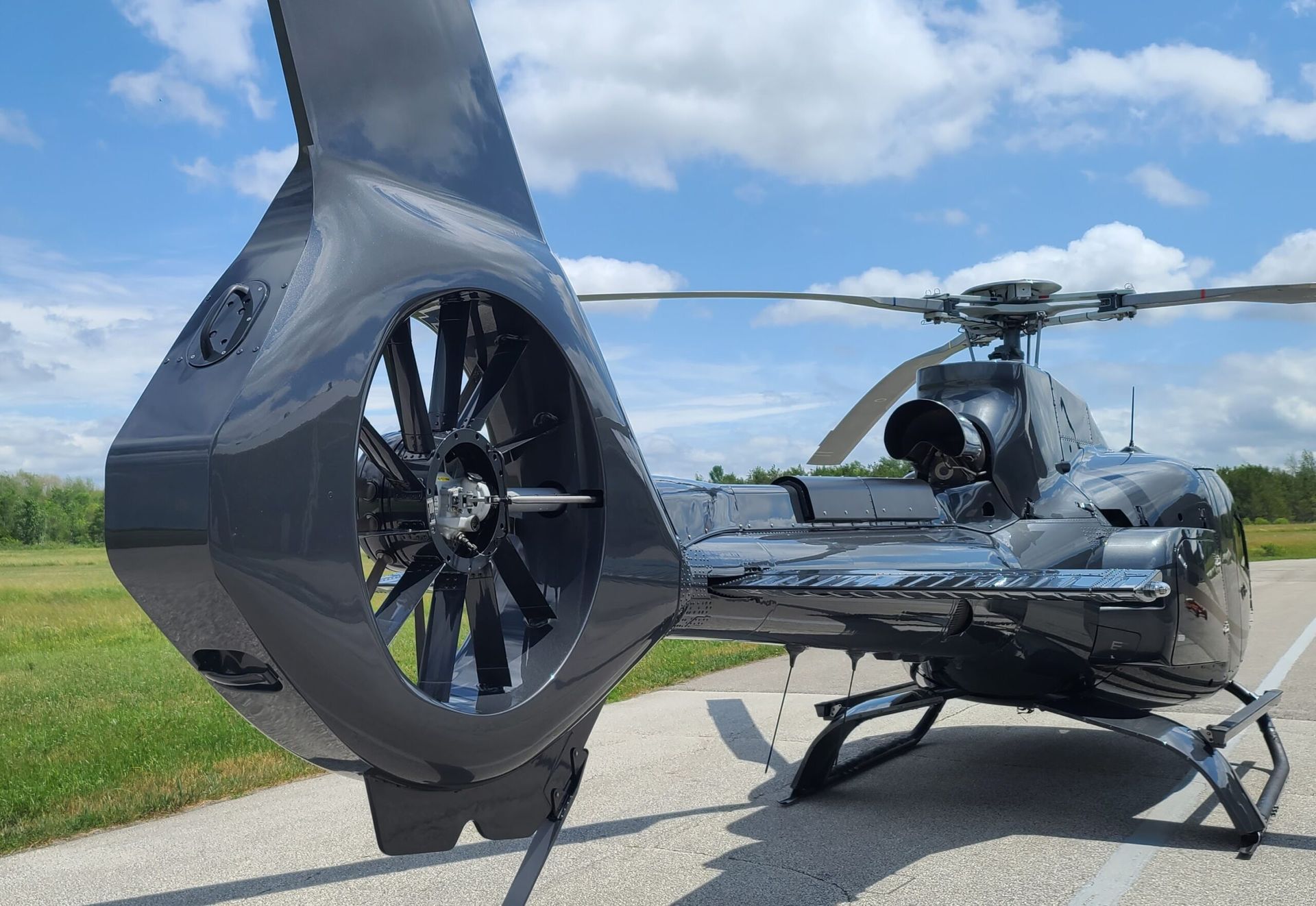 Dark gray helicopter on a runway, tail rotor visible. Blue sky, grass, and trees in the background.