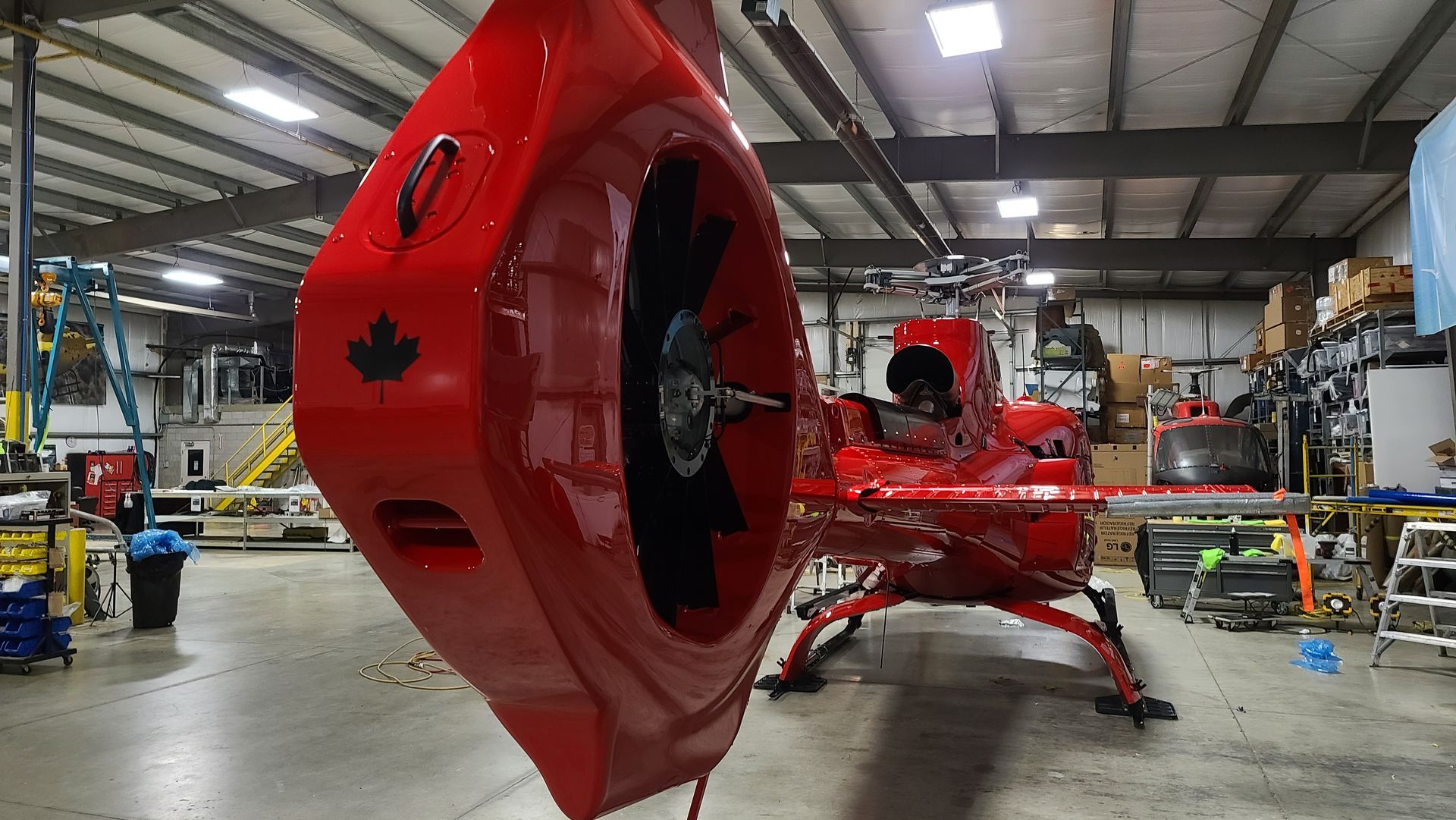 Red helicopter tail section with rotor blades, indoors in a shop setting. Canadian flag symbol visible.