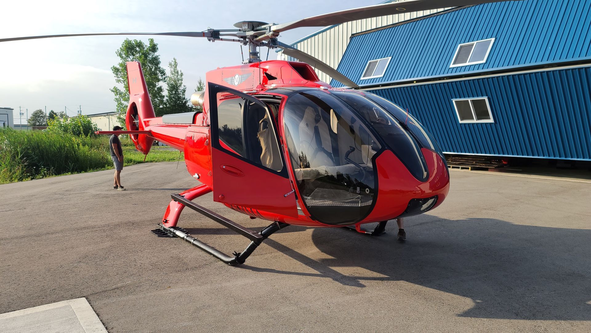 Red helicopter on a paved surface near a blue building; rotor blades are up.
