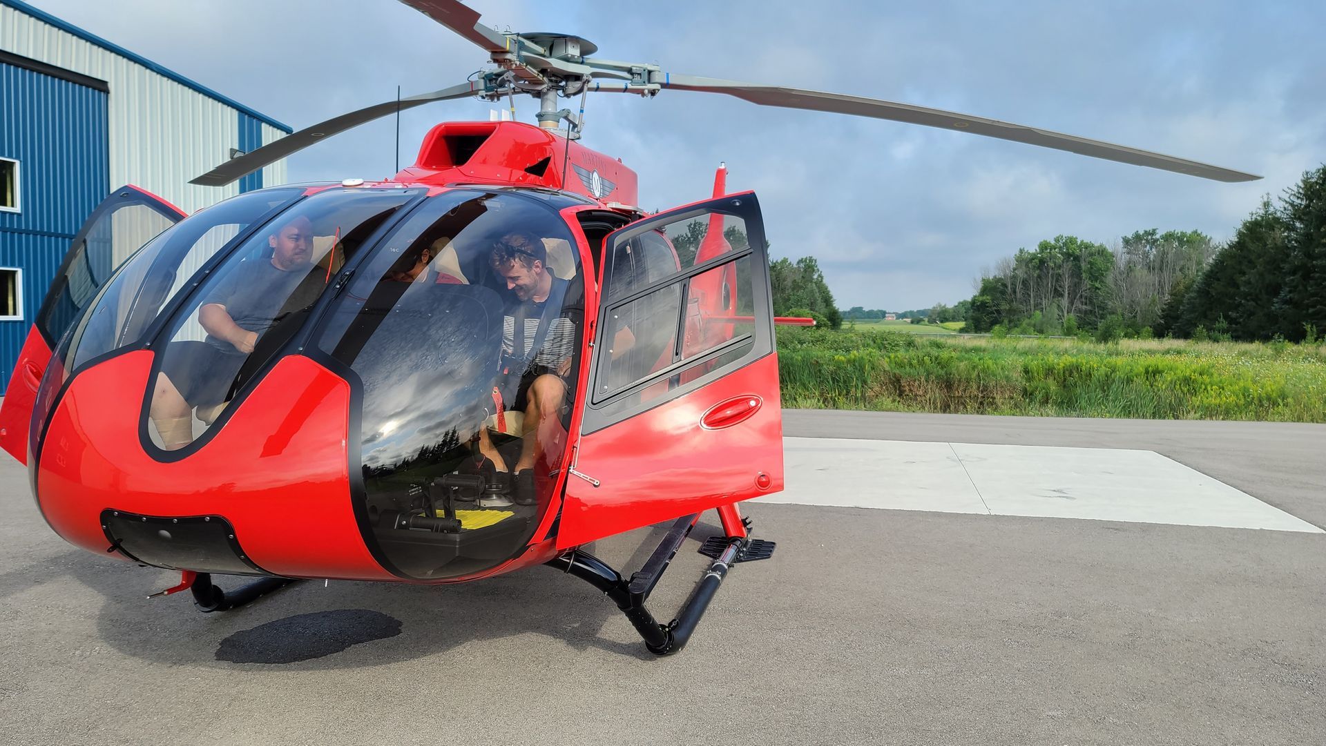 Red helicopter with open doors, two people inside, parked on a pad.