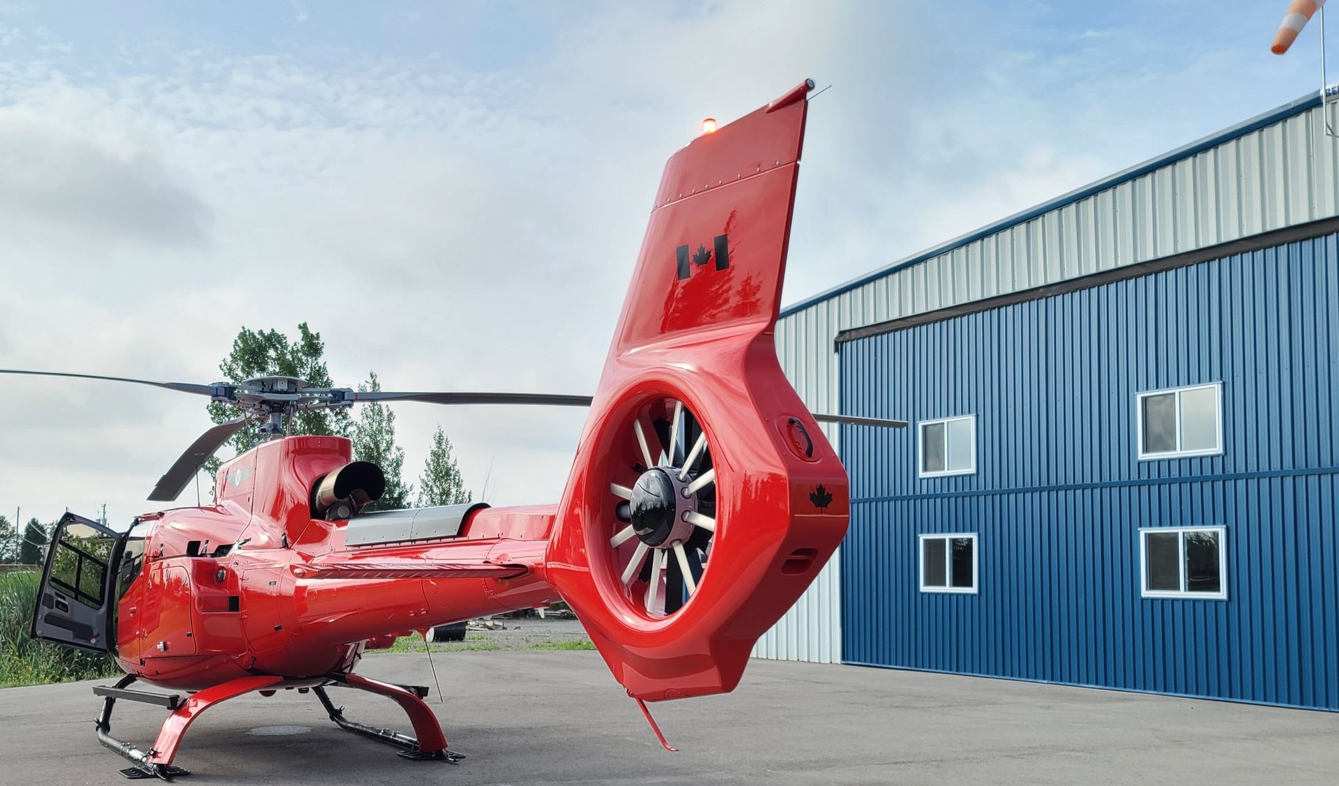 Red helicopter parked near a blue metal building with small windows.