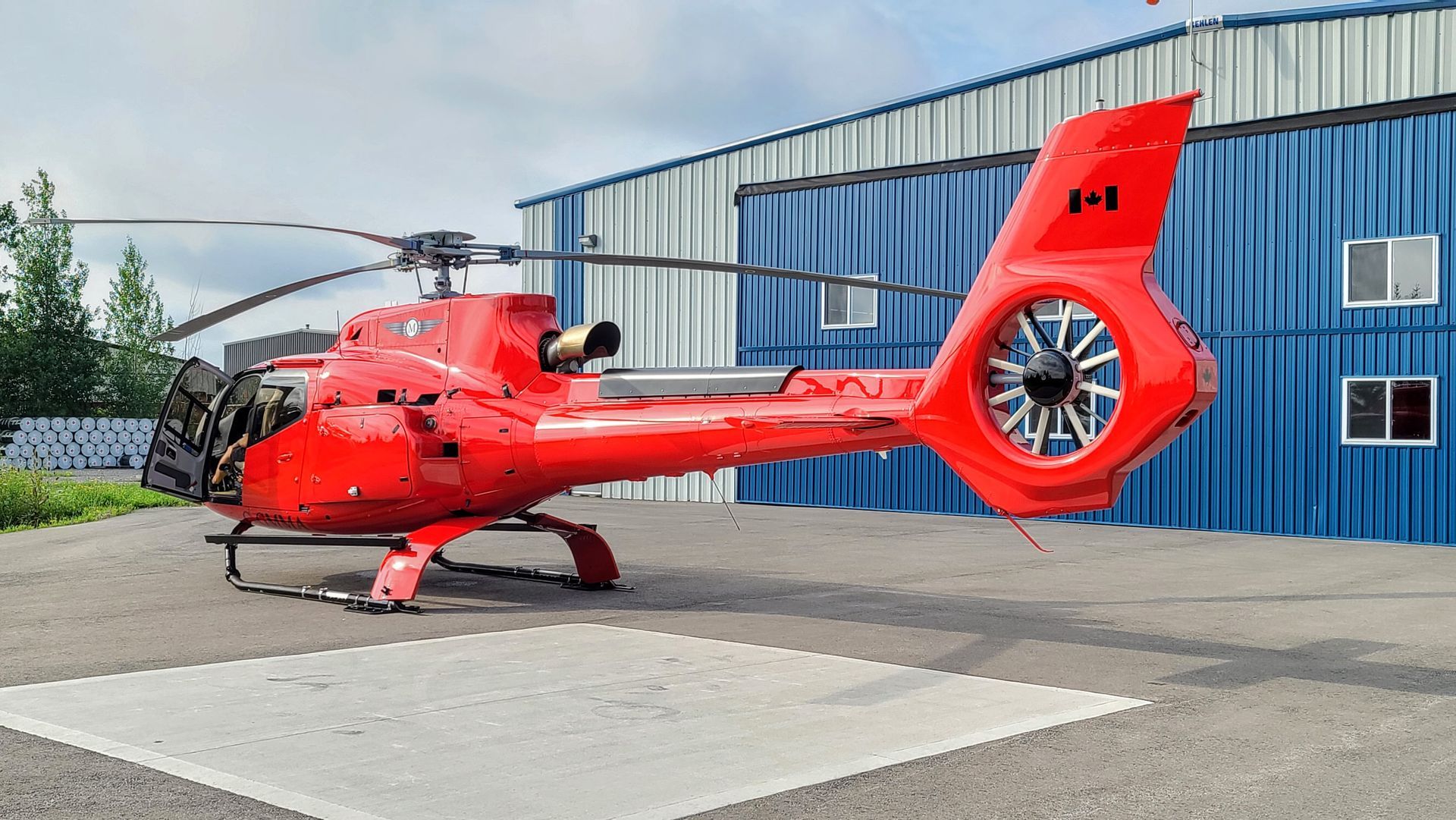 Red helicopter on a gray pad in front of a blue hangar with a Canadian flag on the tail.
