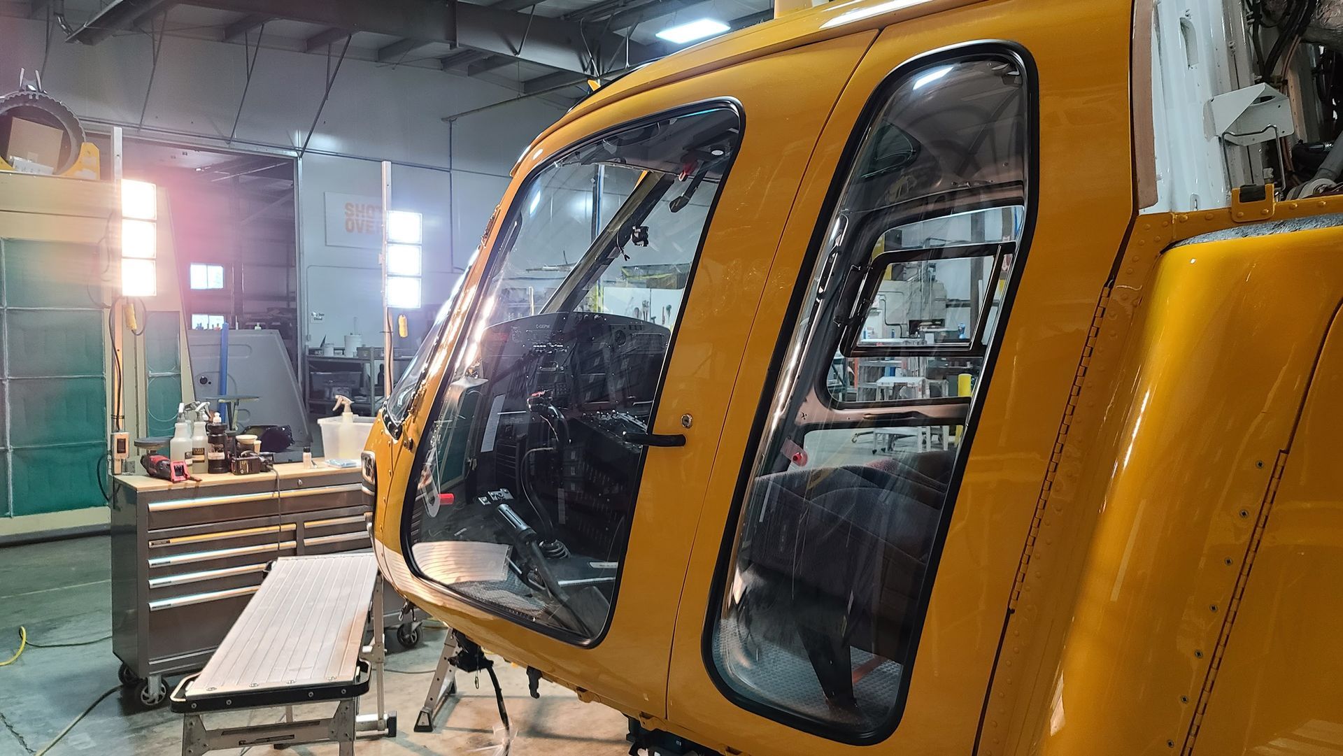 Yellow helicopter cockpit inside a hangar, with open door revealing interior, lit by work lights.