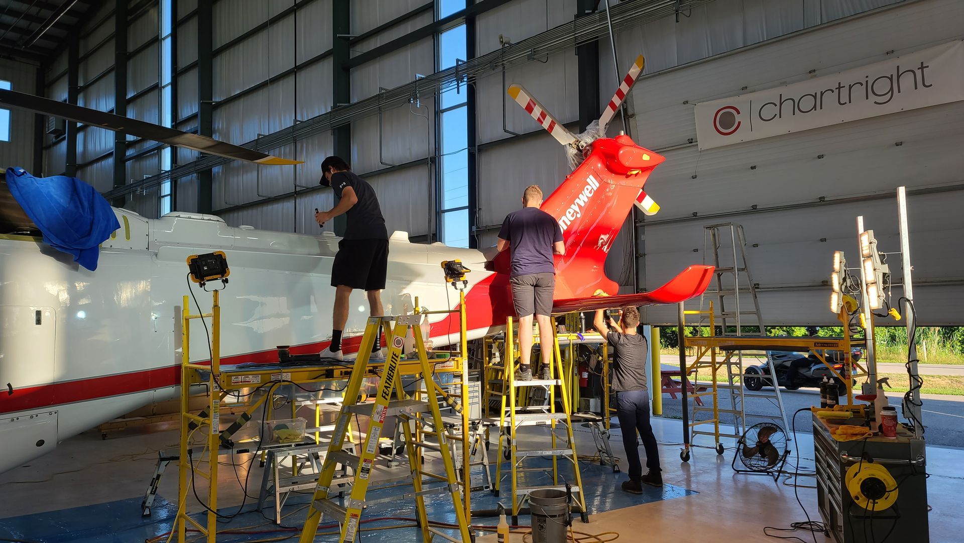 People working on a red and white helicopter in a hangar, using scaffolding.