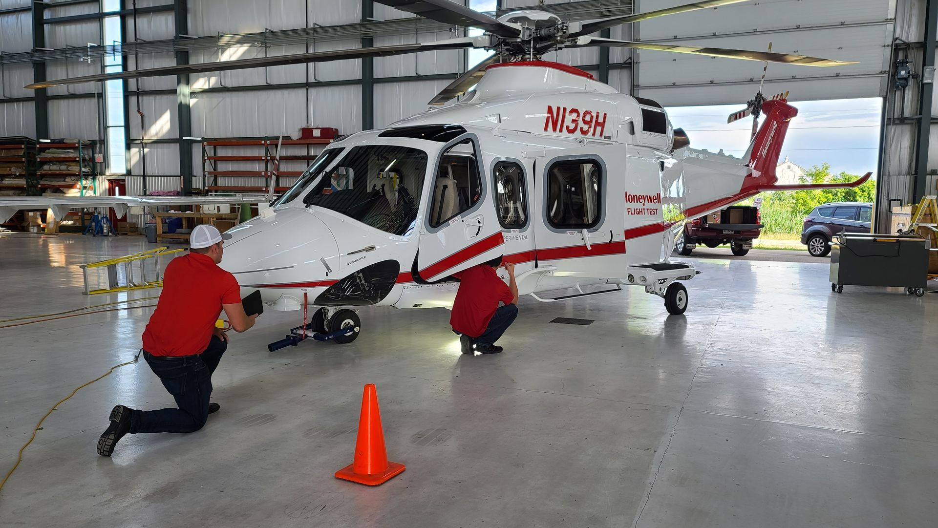 Two people in red shirts inspect a white and red helicopter inside a hangar. Orange cone in front.