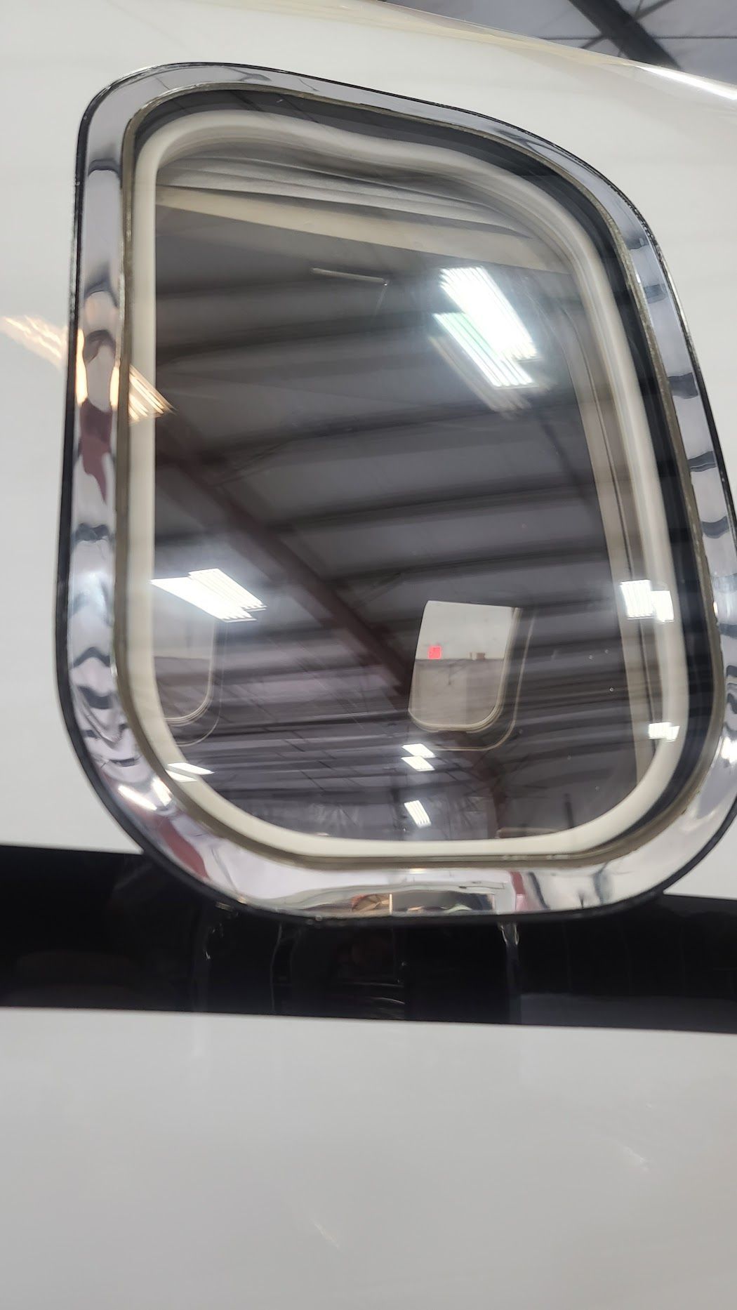Airplane window with a chrome frame reflecting a hangar interior with lights.