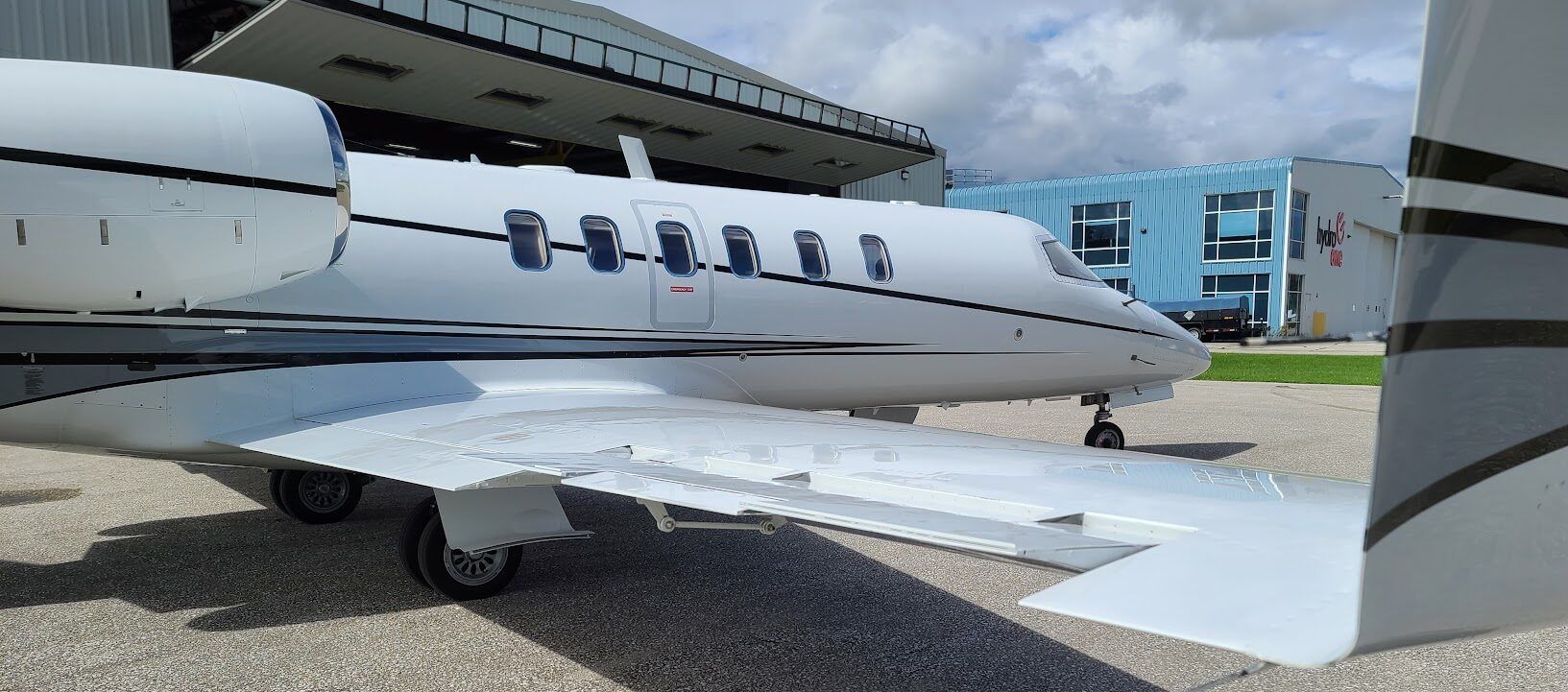 White private jet parked in front of a hangar and blue building, with a wing in the foreground.