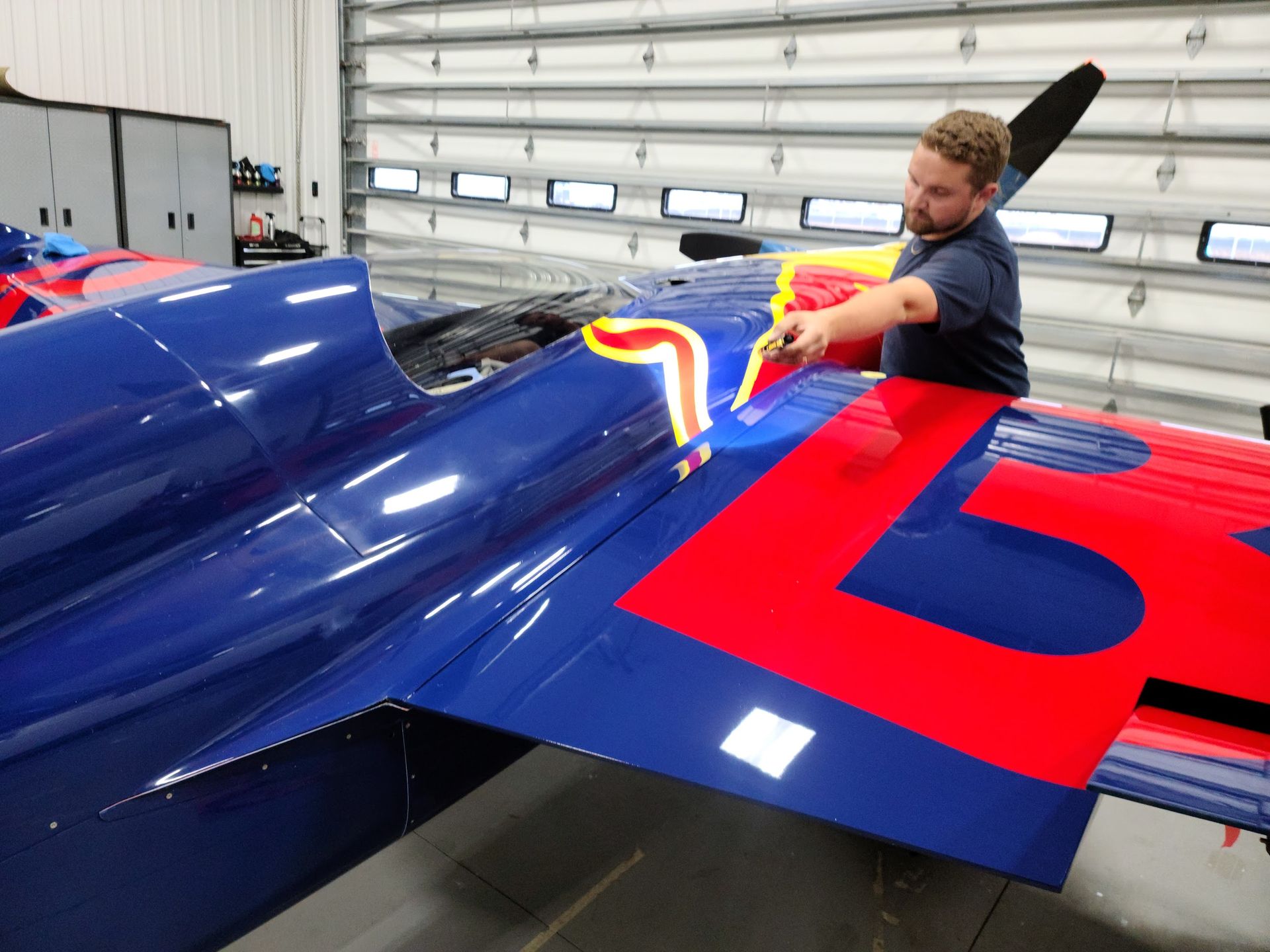 Man working on a bright blue airplane wing with large red numbers in a hangar.