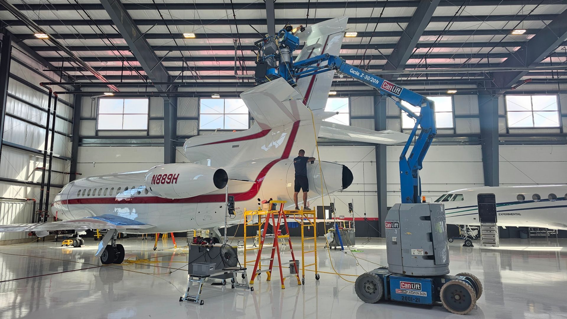 Aircraft maintenance in a hangar; a person on a lift works on the tail; another person on a ladder; equipment on the floor.
