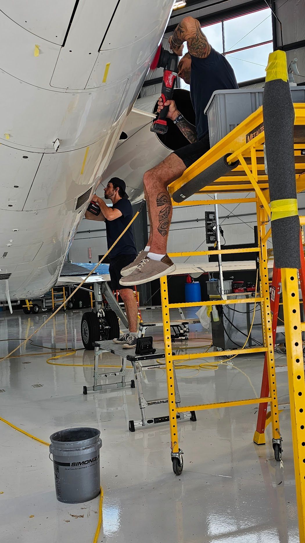 Two workers on scaffolding, repairing aircraft fuselage in a hangar. One uses a drill.