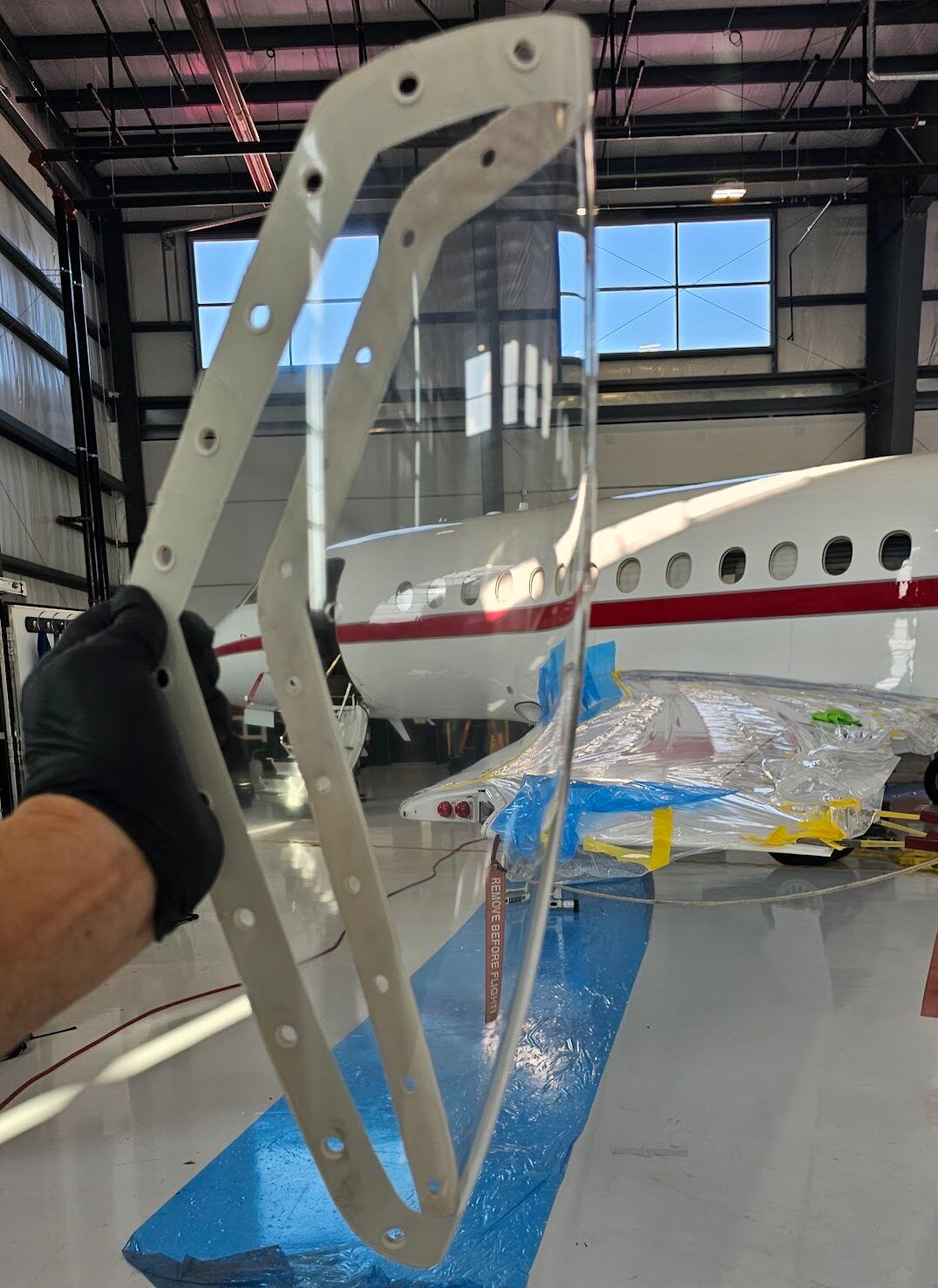 A person holds a clear aircraft window frame with a white border against a hangar backdrop with an airplane.