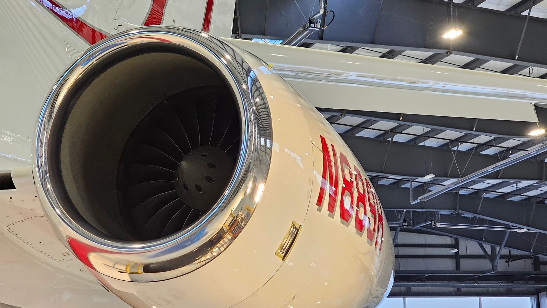 Jet engine, white and red plane exterior; focus on engine intake and exposed fan blades, under hangar lights.