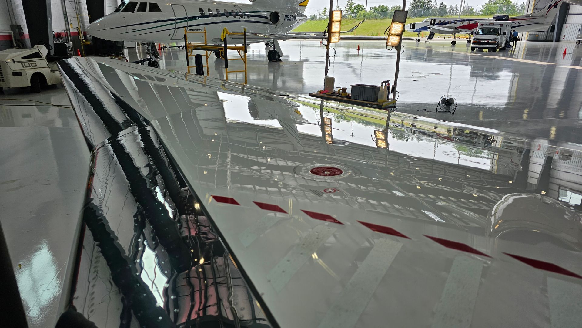 Airplane wing in a hangar, reflecting water and lights. Red stripes on the wing. Other planes and equipment visible.
