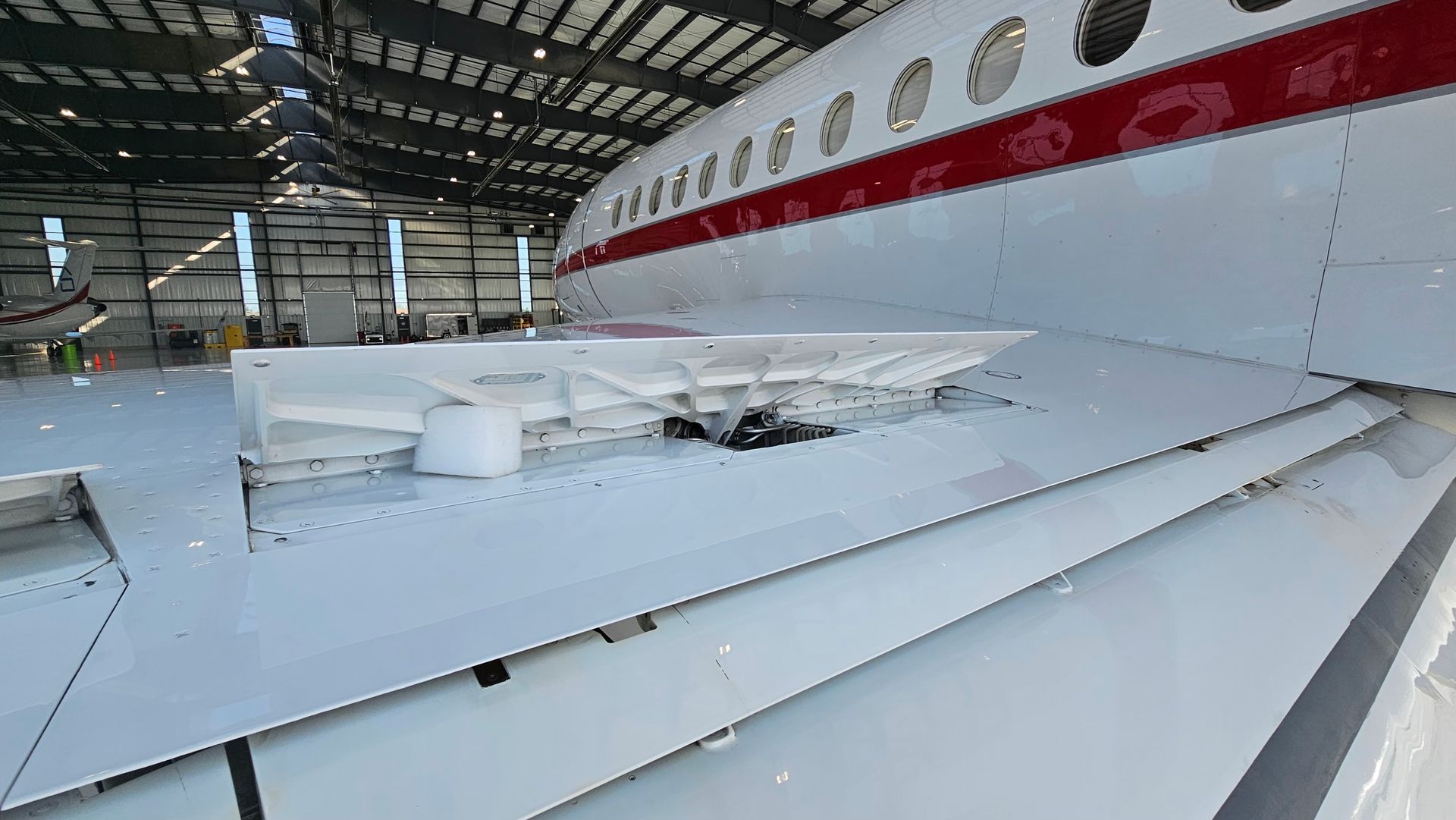 White jet wing with damaged flap in a hangar, red stripe on fuselage.