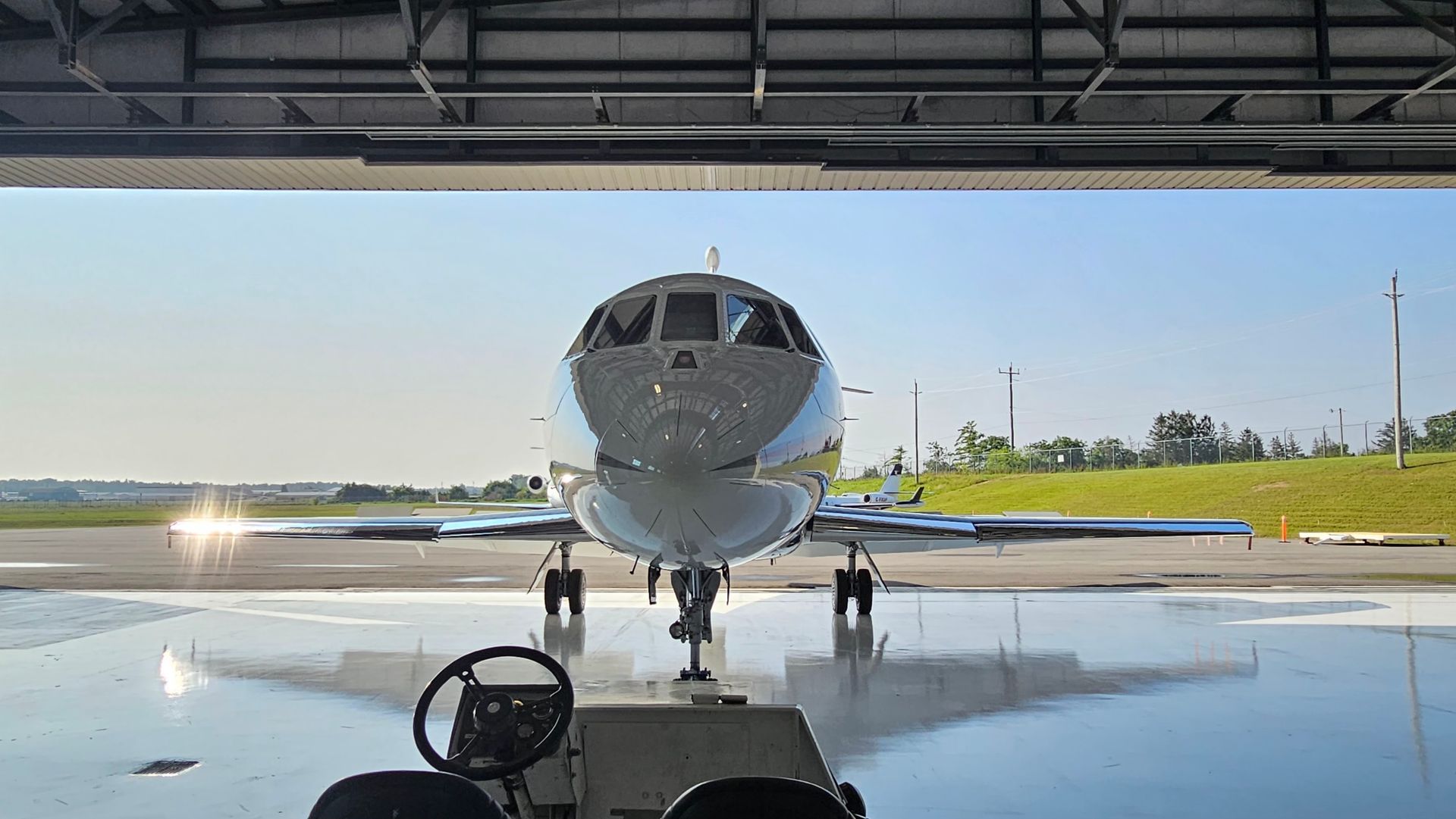 A private jet parked in a hangar, facing the camera. Sunlight reflects off its polished surface.