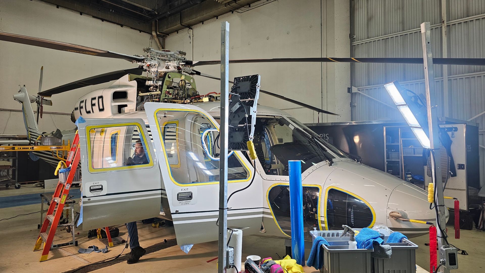 Helicopter being worked on in a hangar. Doors open, maintenance tools visible, with a ladder leaning on the side.