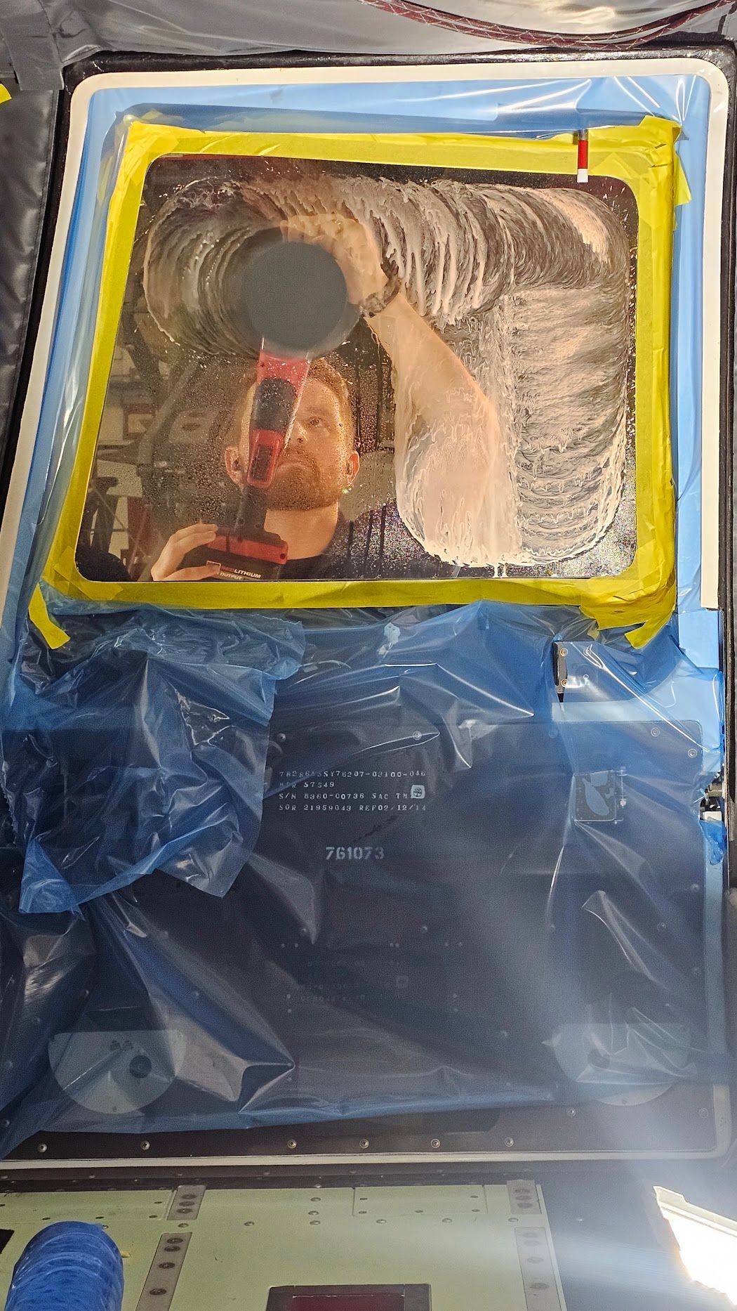 Person wiping soapy film from a taped window. Blue plastic and yellow tape protect the surrounding area.