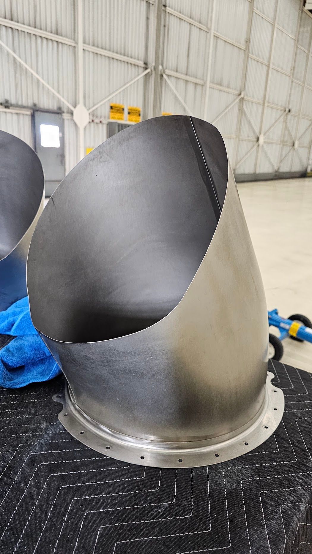 Large, metallic, angled cylindrical component with a bolted base in a hangar setting.