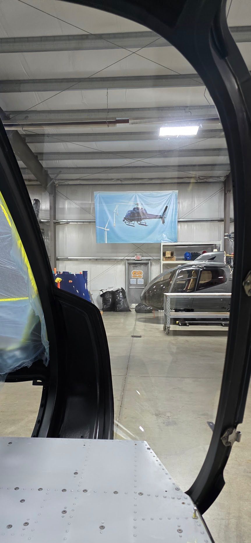 Inside a hangar, a helicopter door frame partially visible; poster with a helicopter image is hung on the wall.