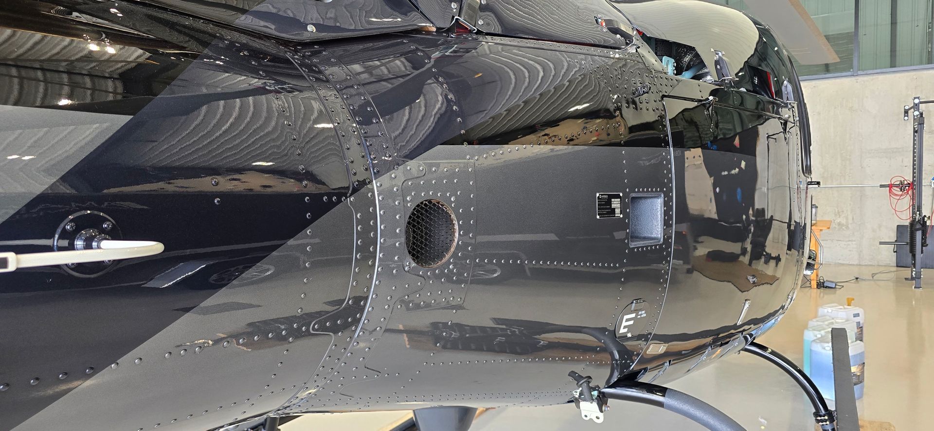 Close-up of a black helicopter fuselage in a hangar. A circular opening and other details are visible.