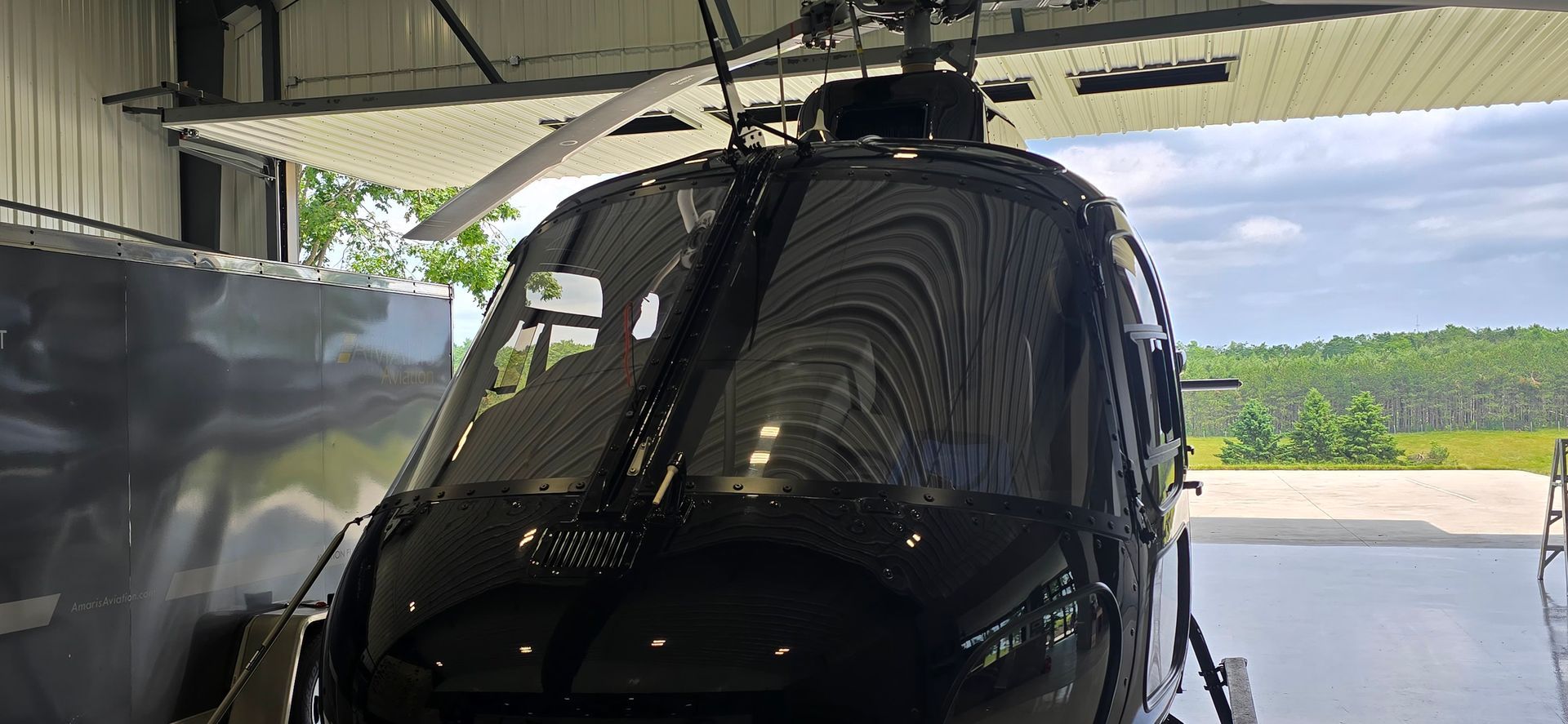 Black helicopter inside a hangar, with blades extended. Green trees and cloudy sky visible in the background.