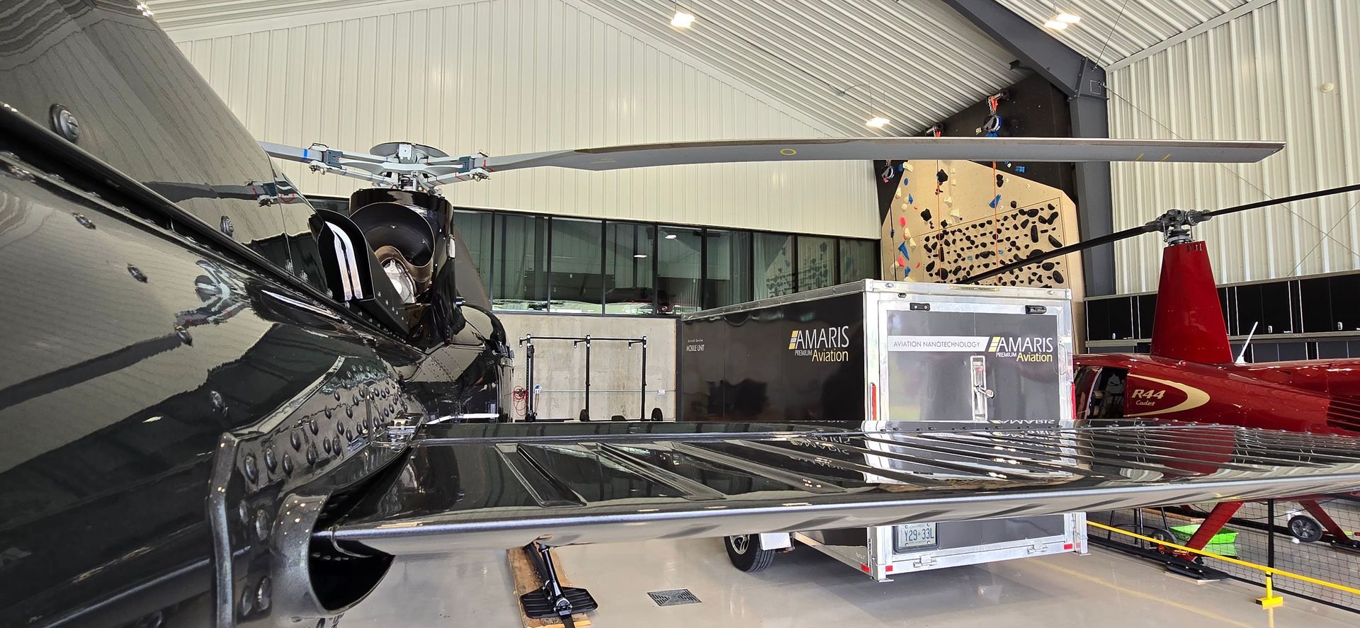Helicopter in a hangar; rotor blades extended. Black and red aircraft. Silver trailer in foreground.