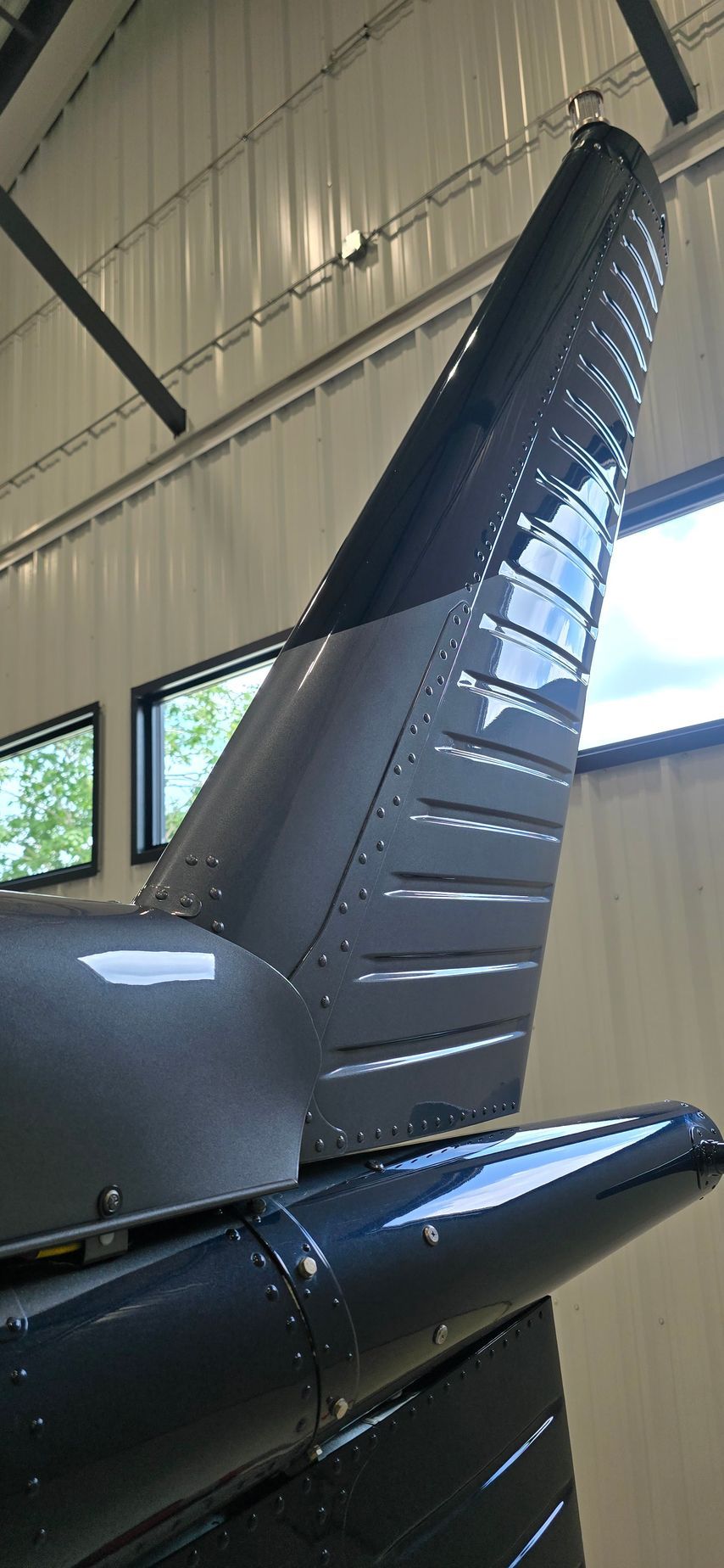 Tail of a dark-colored aircraft in a hangar. Features include vertical stabilizer, rivets, and reflective surfaces.