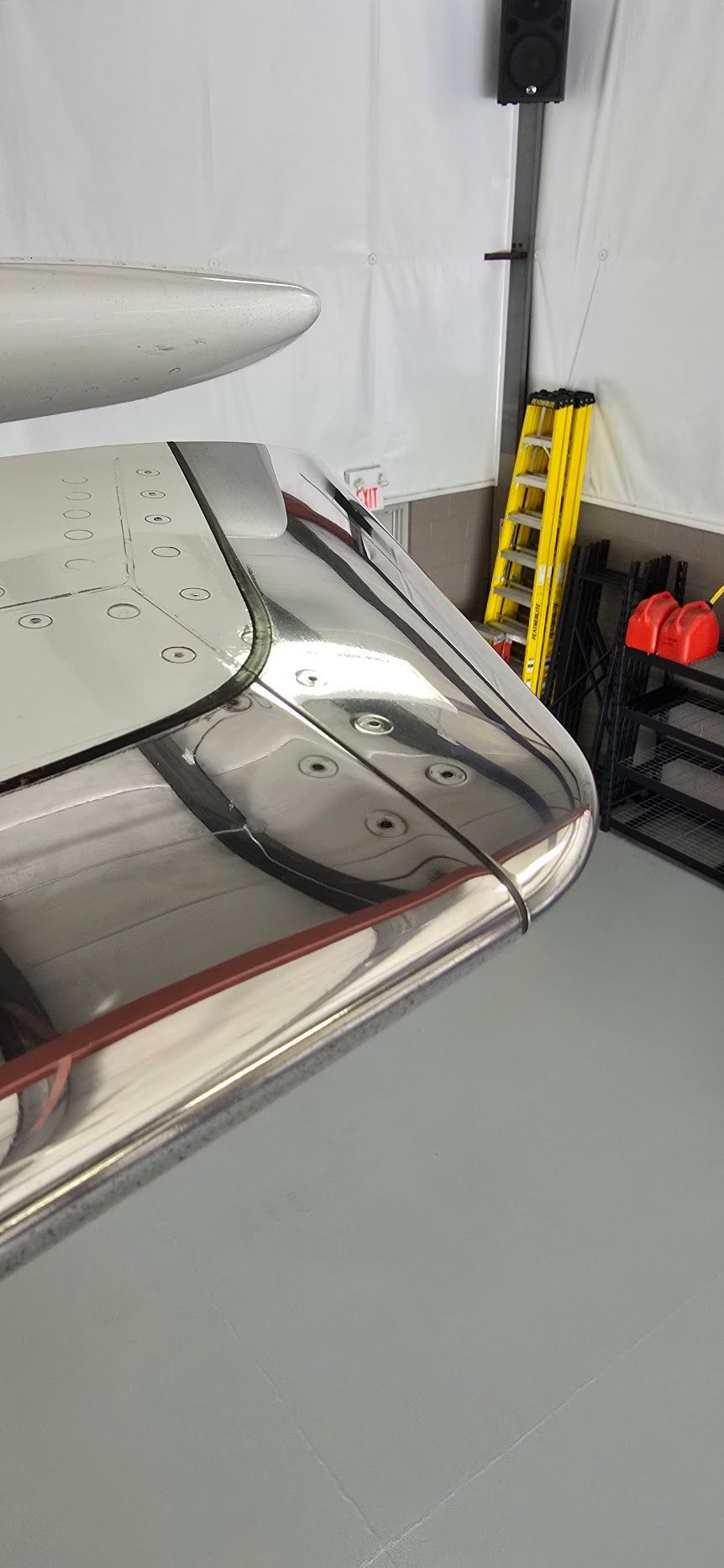 Close-up view of a polished metal aircraft wing with rivets, in a hangar, with a ladder and a storage rack.