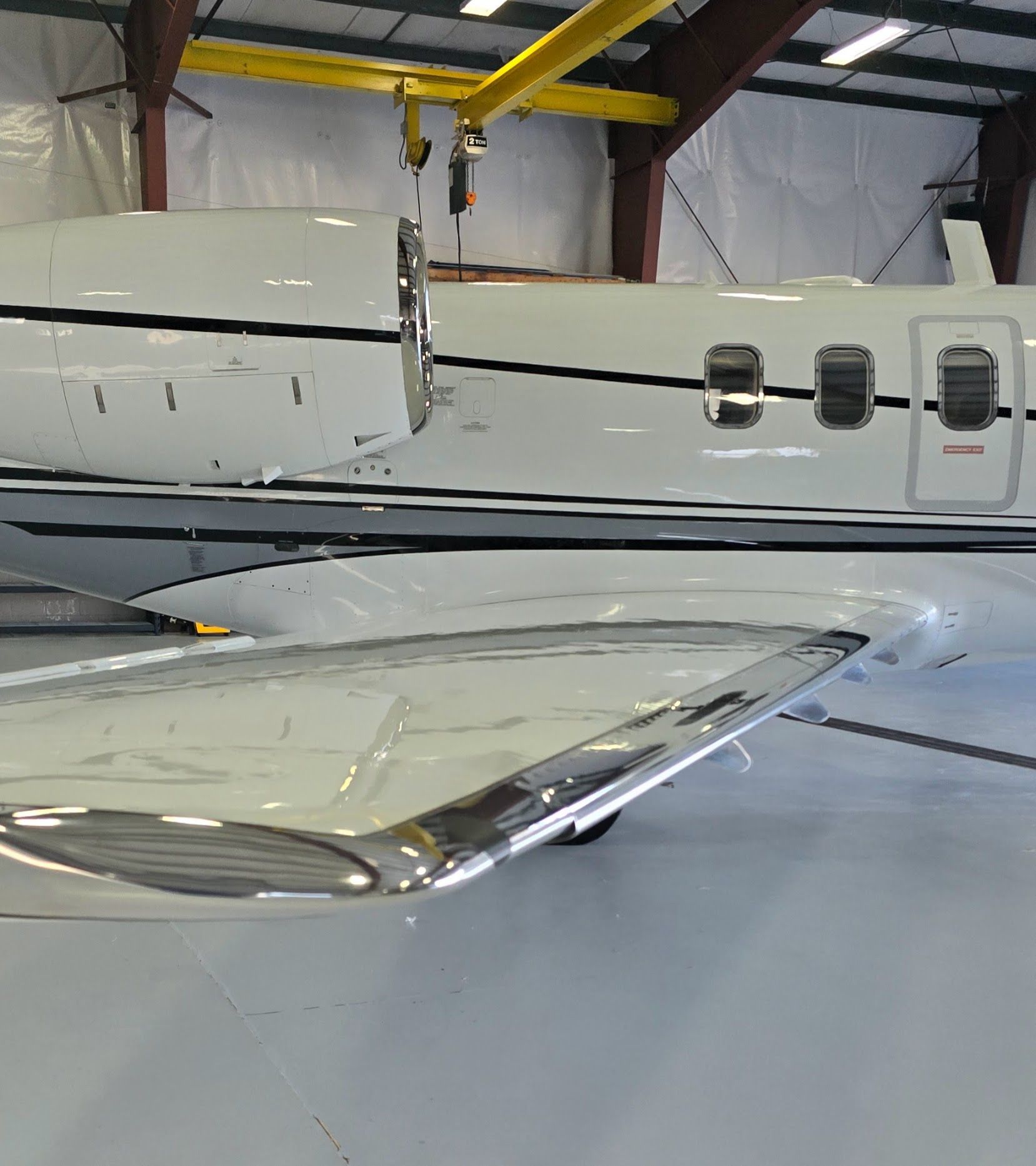 White private jet wing and fuselage in a hangar, winglet visible, overhead crane.