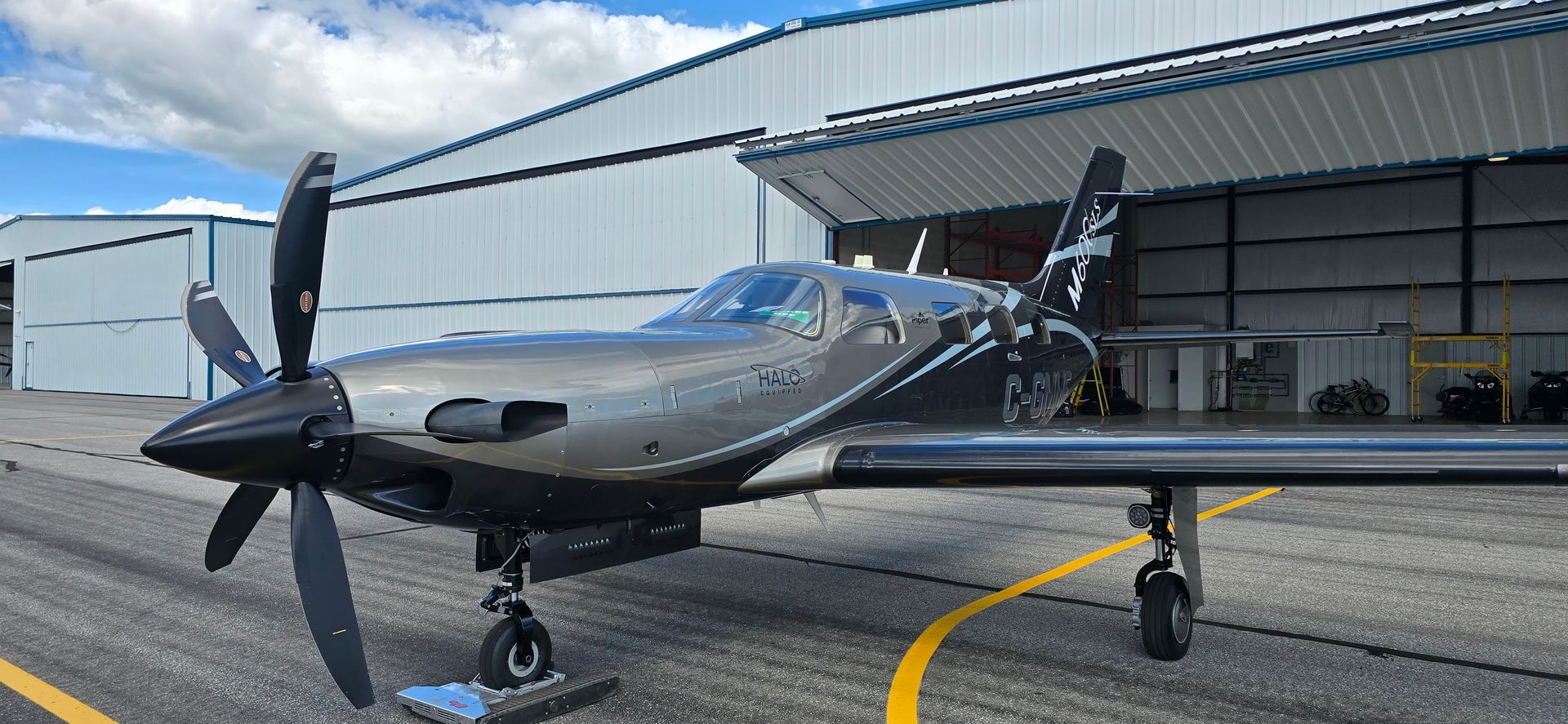 A gray turboprop airplane parked on a tarmac in front of a hangar under a blue sky.