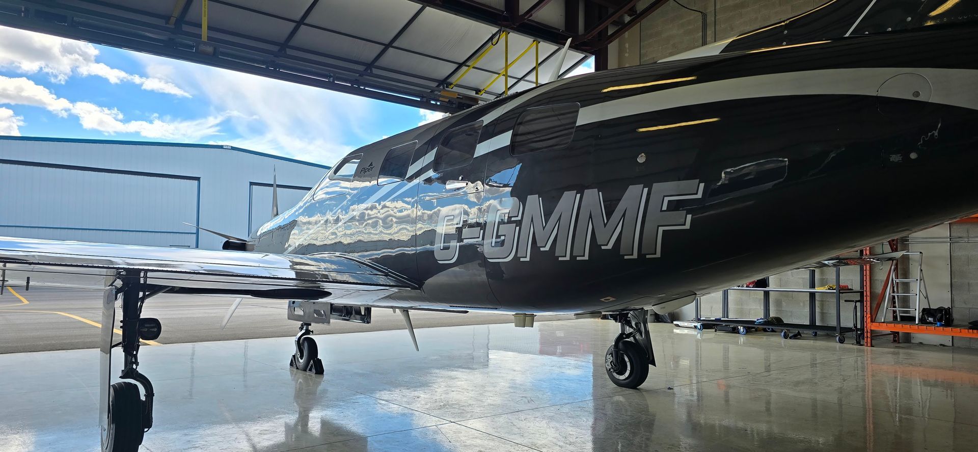 Black airplane in a hangar; registration code G-GMMF visible. Bright reflections on the wet floor.