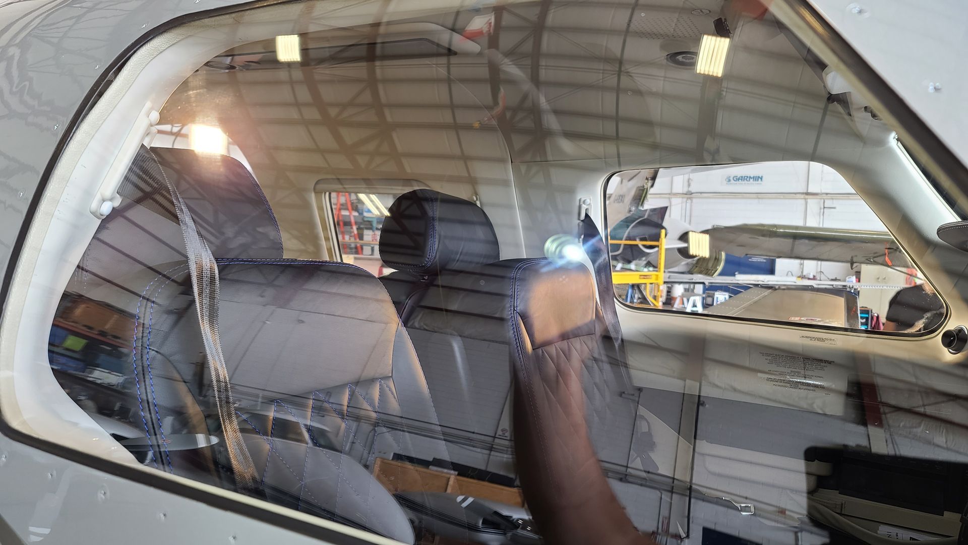 Inside an airplane cockpit, a hand touches the window. Airplane seats and hangar in the background.