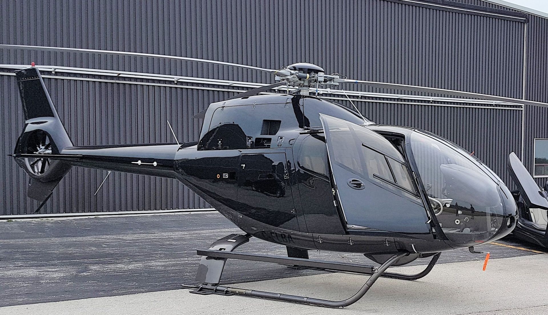 Black helicopter with open door on a concrete surface, parked in front of a building.