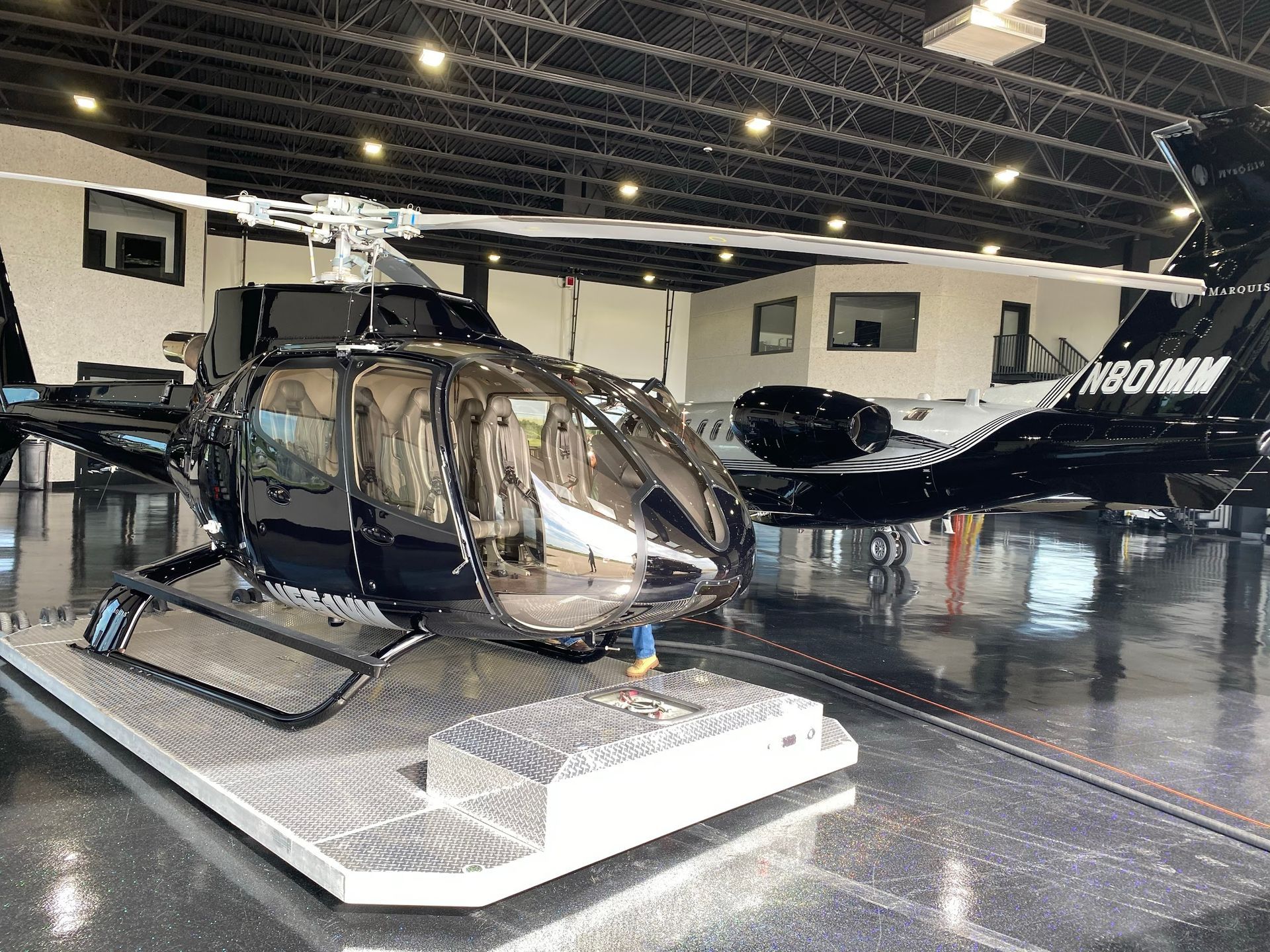 Black helicopter on a metal platform inside a hangar, with a black and white airplane in the background.