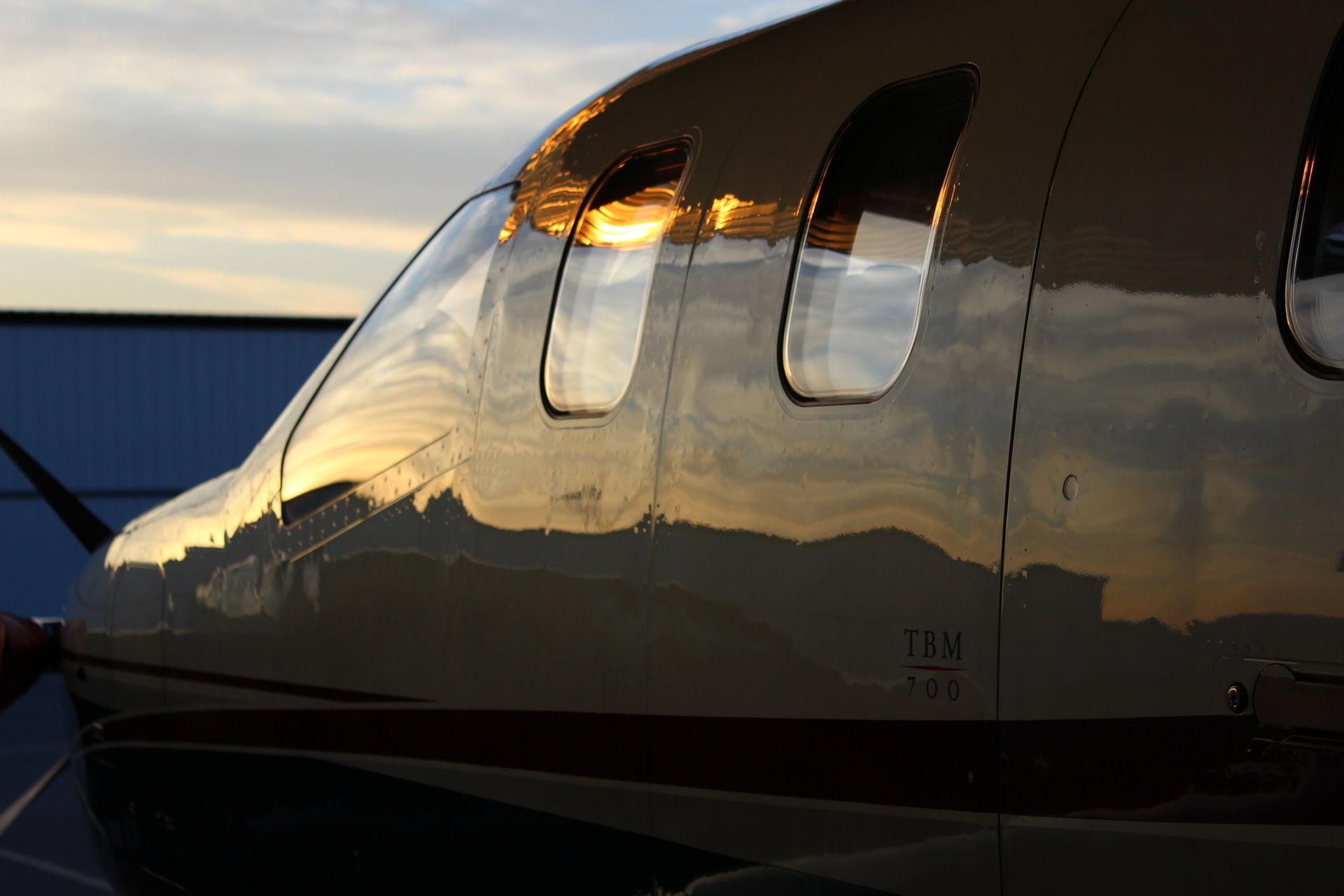 Close-up of a small plane's fuselage reflecting a sunset, with a curved window and the plane's name visible.