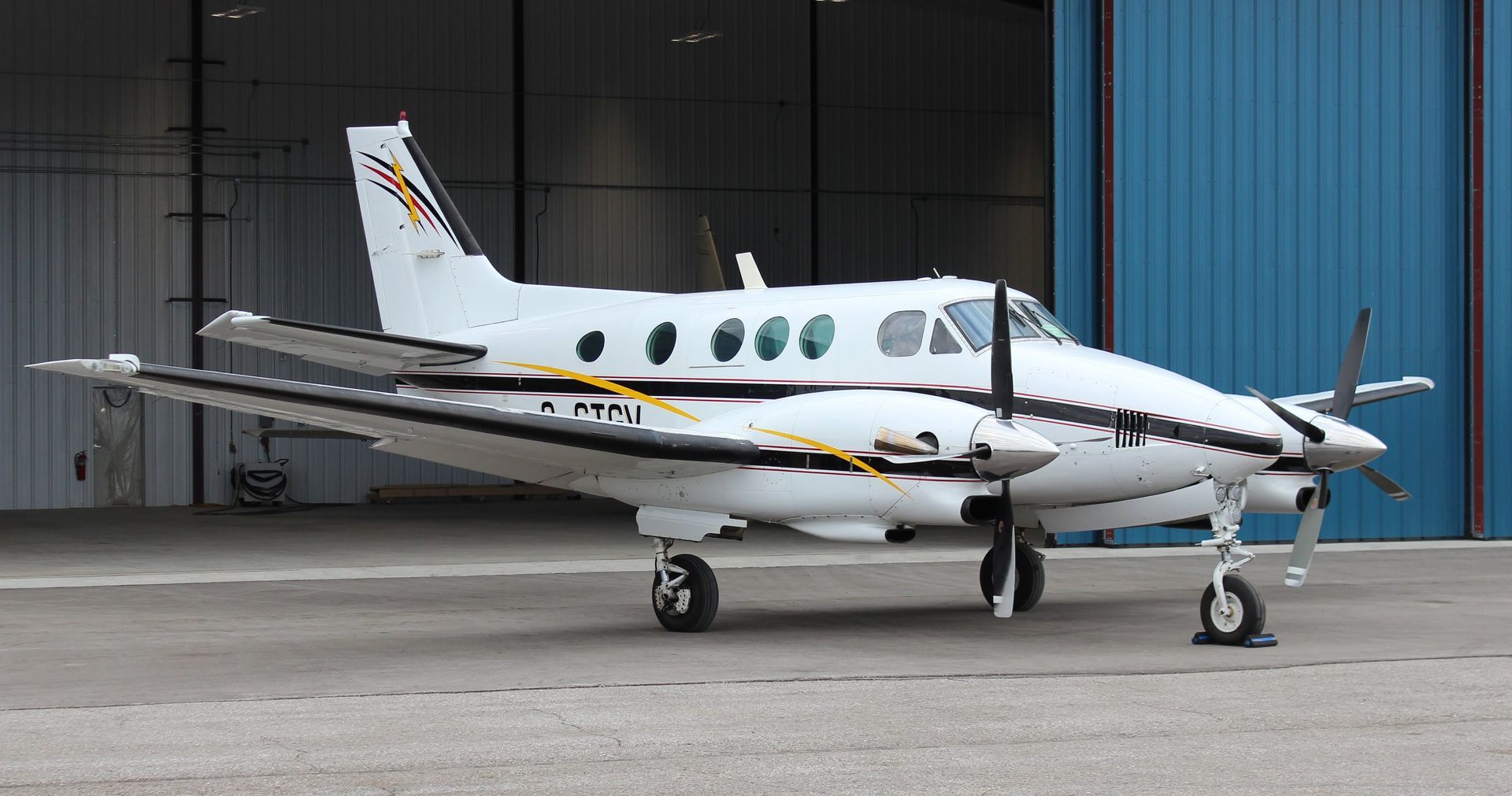 White and black twin-engine airplane parked outside a blue hangar with its propellers at rest.