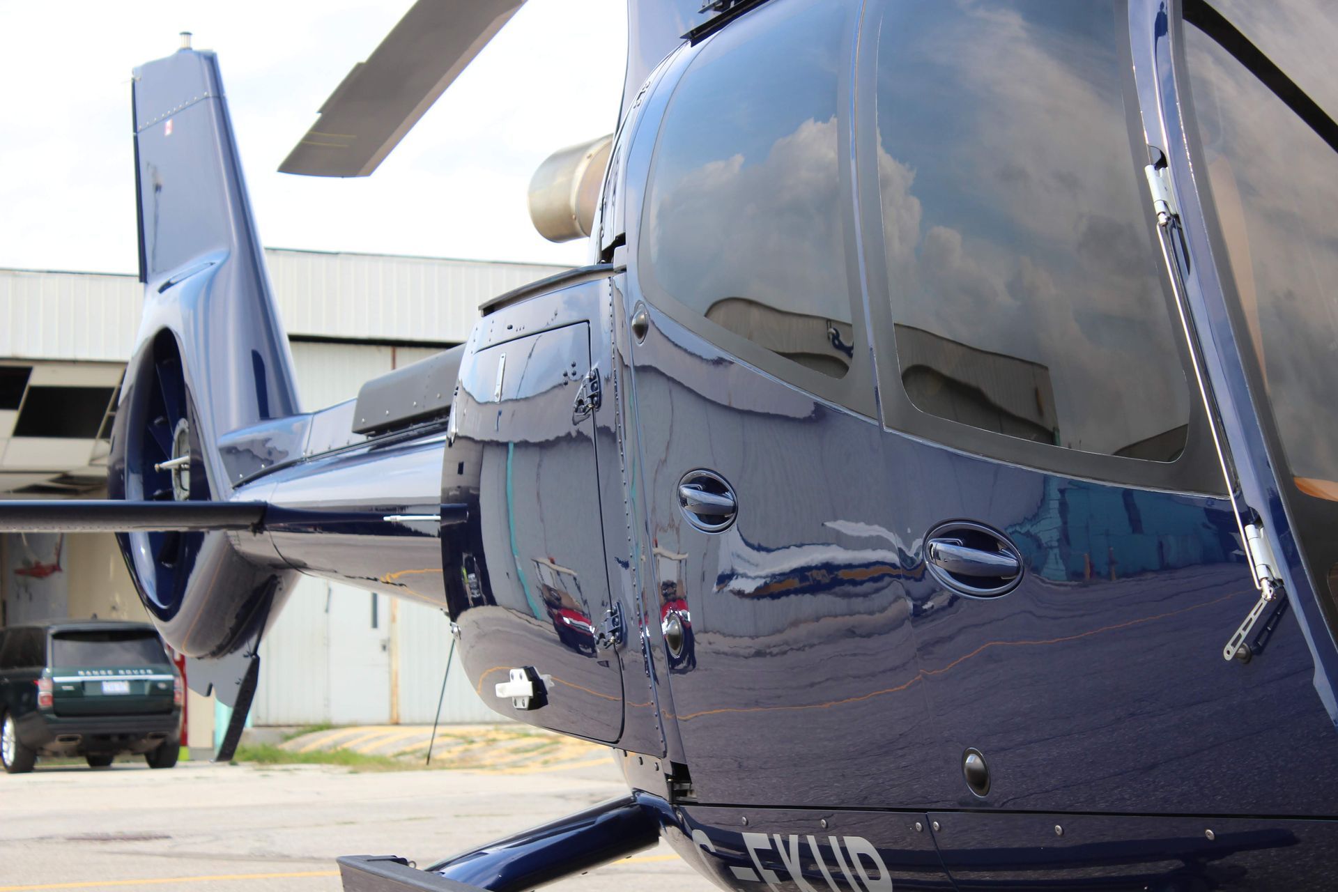 Blue helicopter parked on a tarmac, with a reflection of a building and a vehicle.