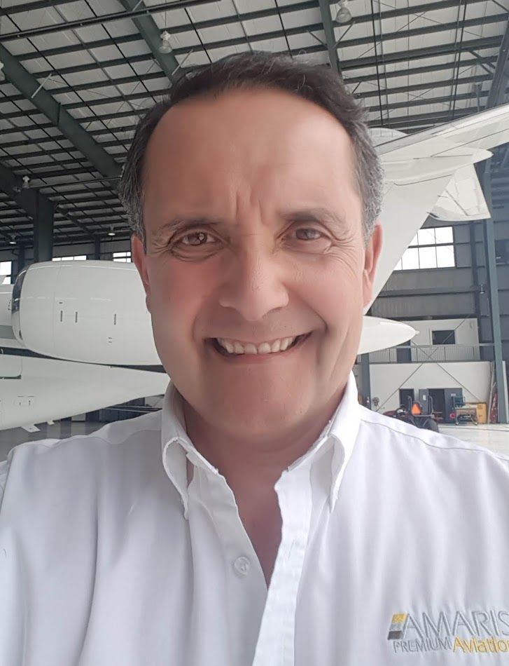 Man smiling in a white shirt, in front of an aircraft in a hangar.