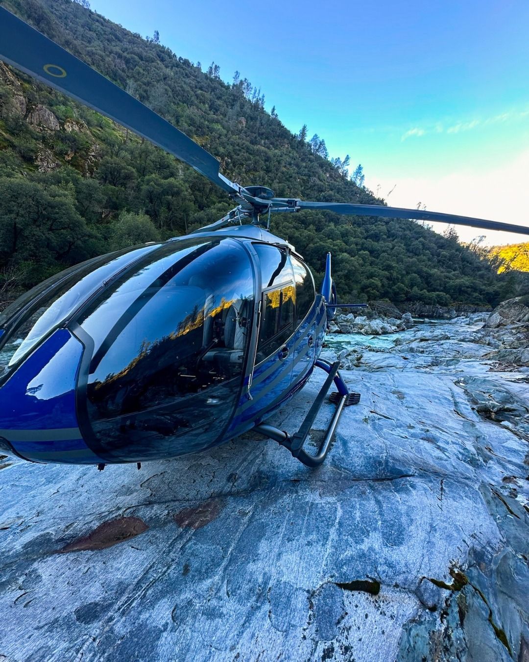 Blue helicopter parked on a rocky riverbed, trees on hillside in the background.
