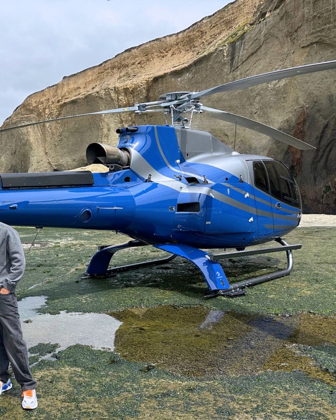 Blue helicopter on a rocky beach near a cliff; a person stands nearby.