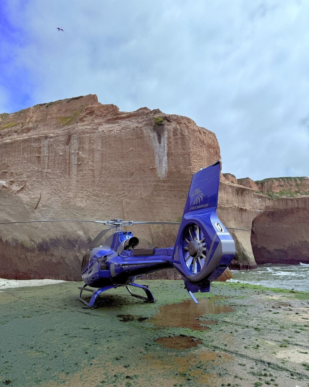 Blue helicopter parked on a beach, near a cliff, under a cloudy sky.