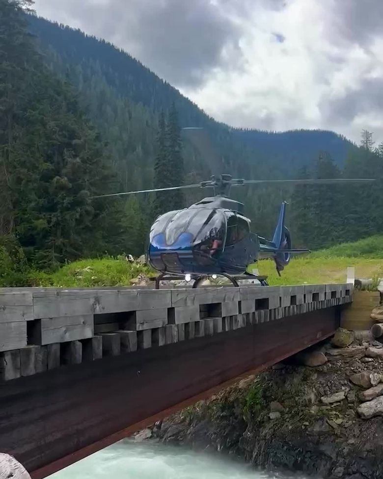 Helicopter lands on a bridge over a river in a mountainous, wooded area.