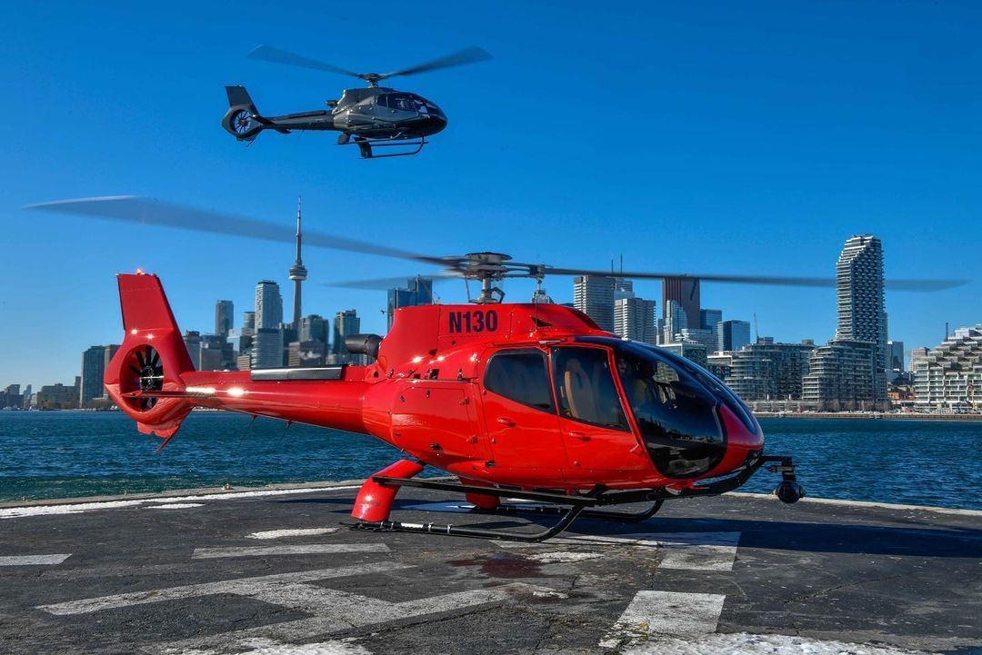 Red helicopter on a helipad with a second helicopter in the sky, Toronto skyline in the background.