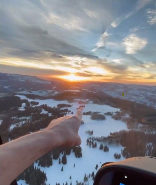 Person pointing at a snow-covered landscape at sunset from a high vantage point, possibly an aircraft.