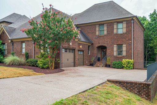 A large brick house with a large driveway and a tree in front of it.