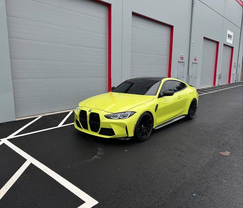 Yellow BMW M4 coupe parked in front of gray industrial building with red trim.